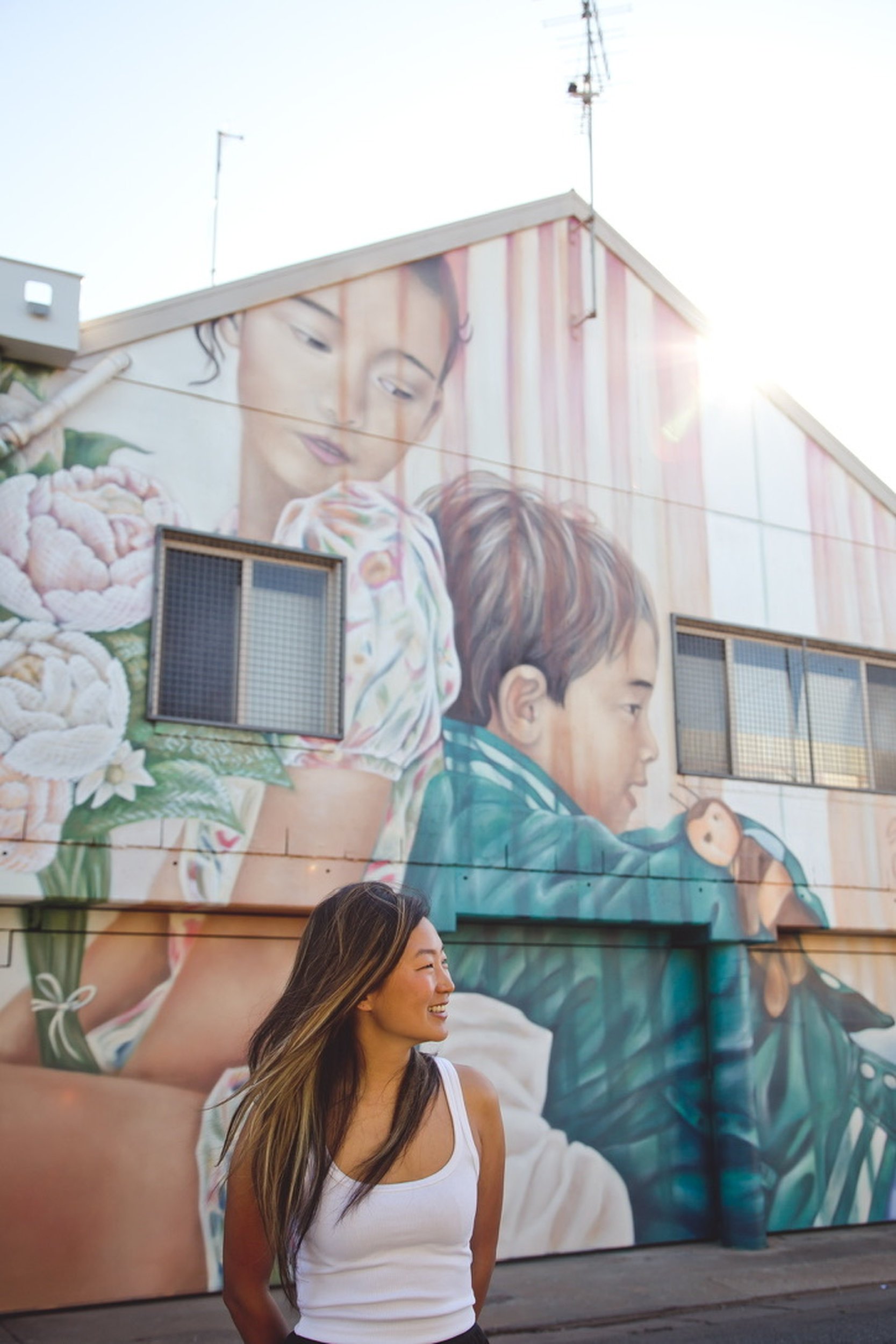 A young woman with long brown hair smiling and looking to the side, standing in front of a colorful mural on a building wall. The mural features large portraits of a woman and a young boy with detailed facial features and vibrant clothing. The photograph is taken during the daytime with sunlight brightening the scene.