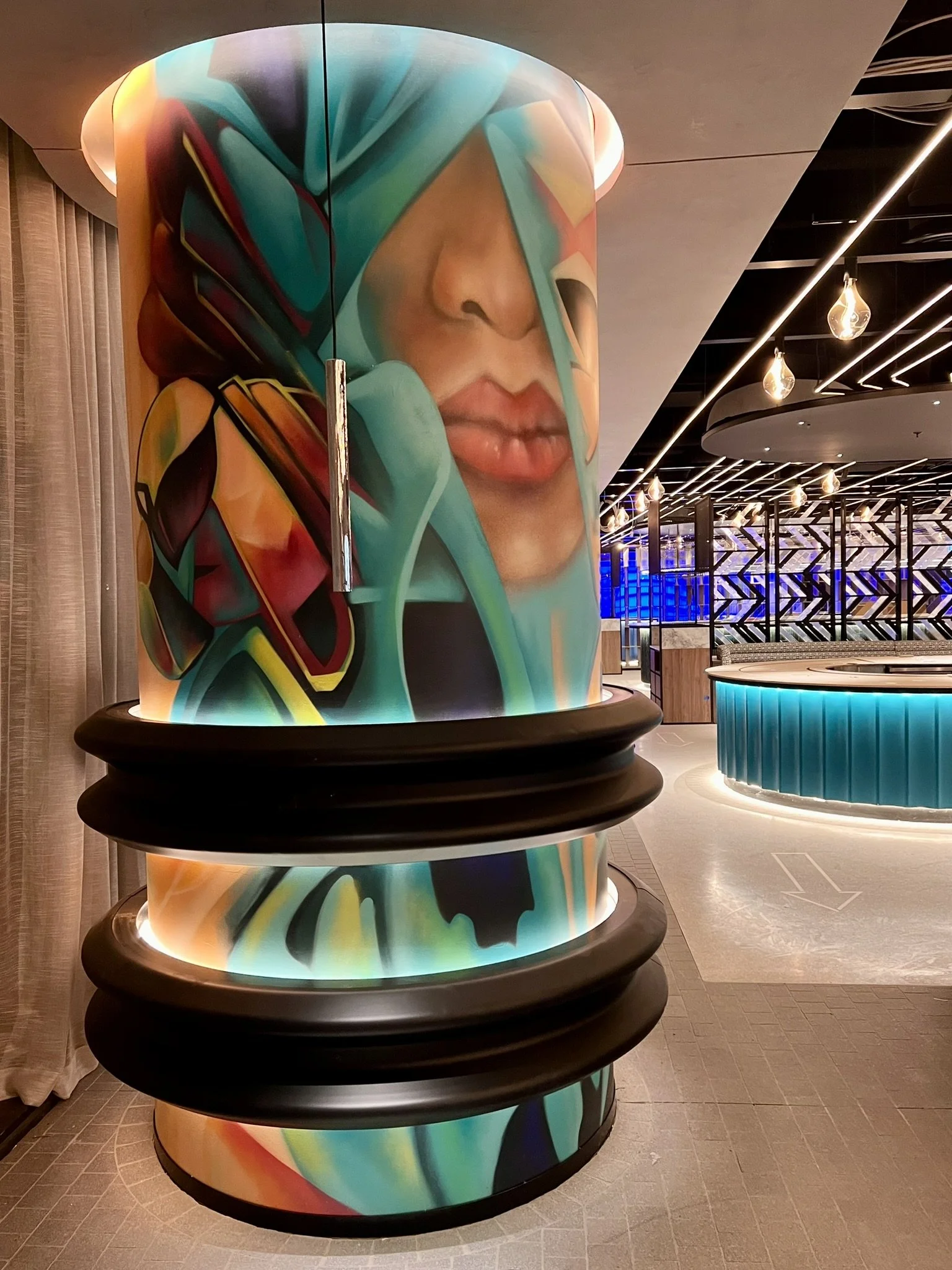 A large cylindrical column with colorful abstract artwork of a woman's face on it, located in a modern indoor public space with ambient lighting and decorative ceiling fixtures.