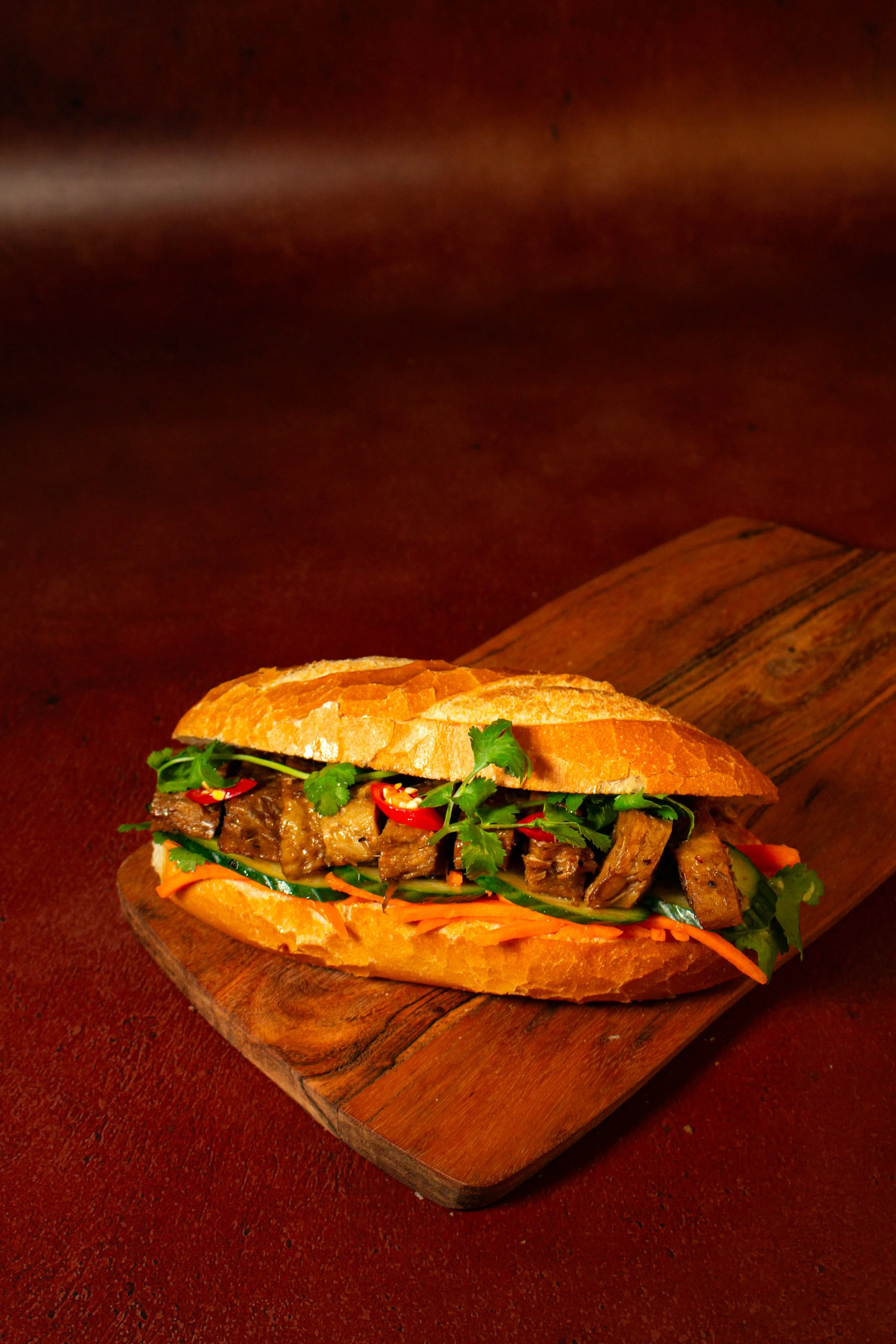 A sandwich made with a crusty baguette, filled with sliced beef, fresh cilantro, red chili slices, cucumber, and shredded carrots, on a wooden serving board.