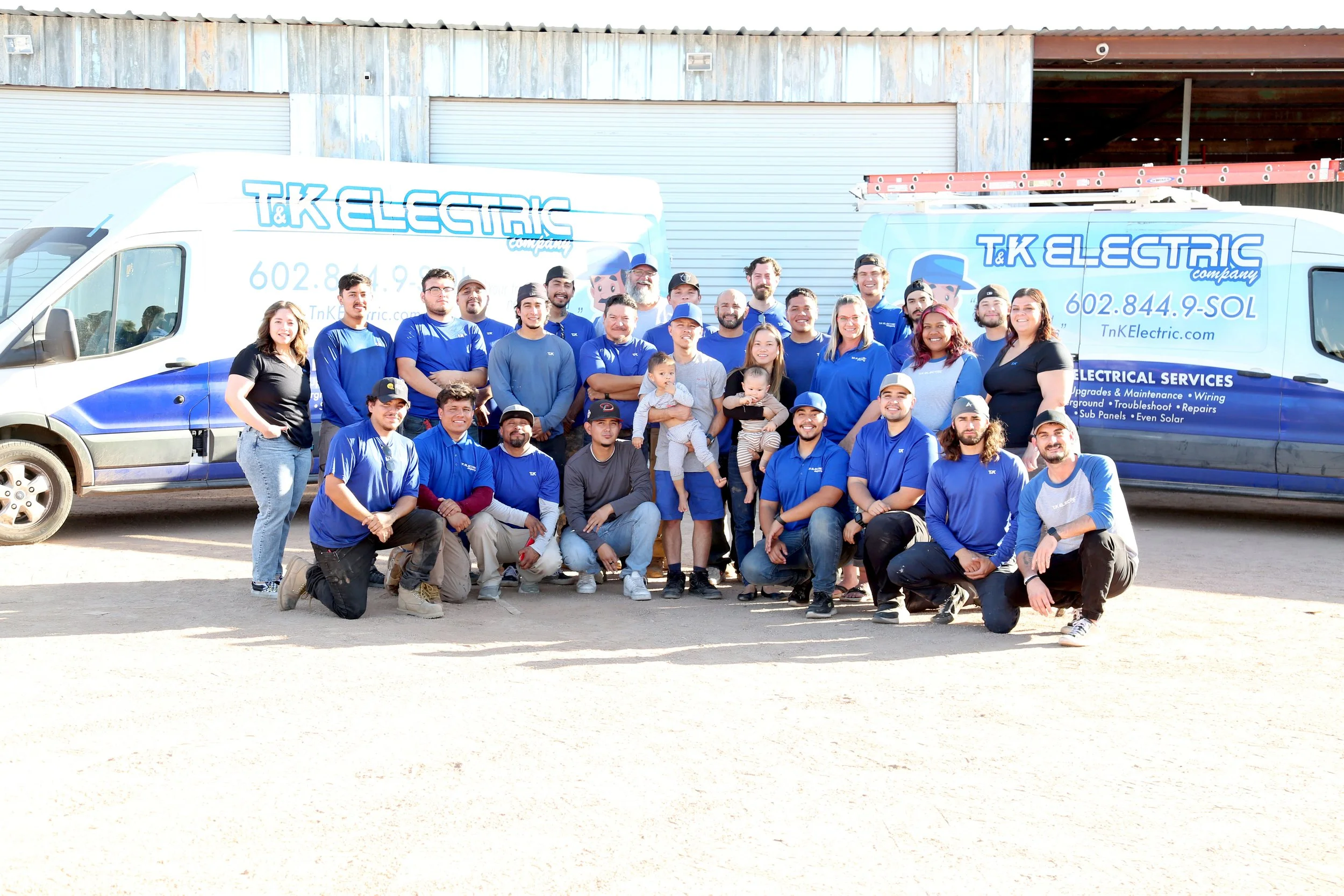 Group of people posing in front of two company service vans with the logo "T&K Electric" during daytime, with some individuals holding children, in front of a building. All are dressed casually, with most wearing blue shirts.