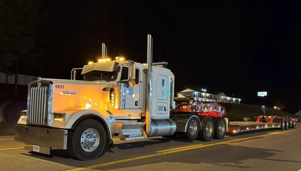 Yellow and white heavy-haul truck with a lowboy trailer at night.