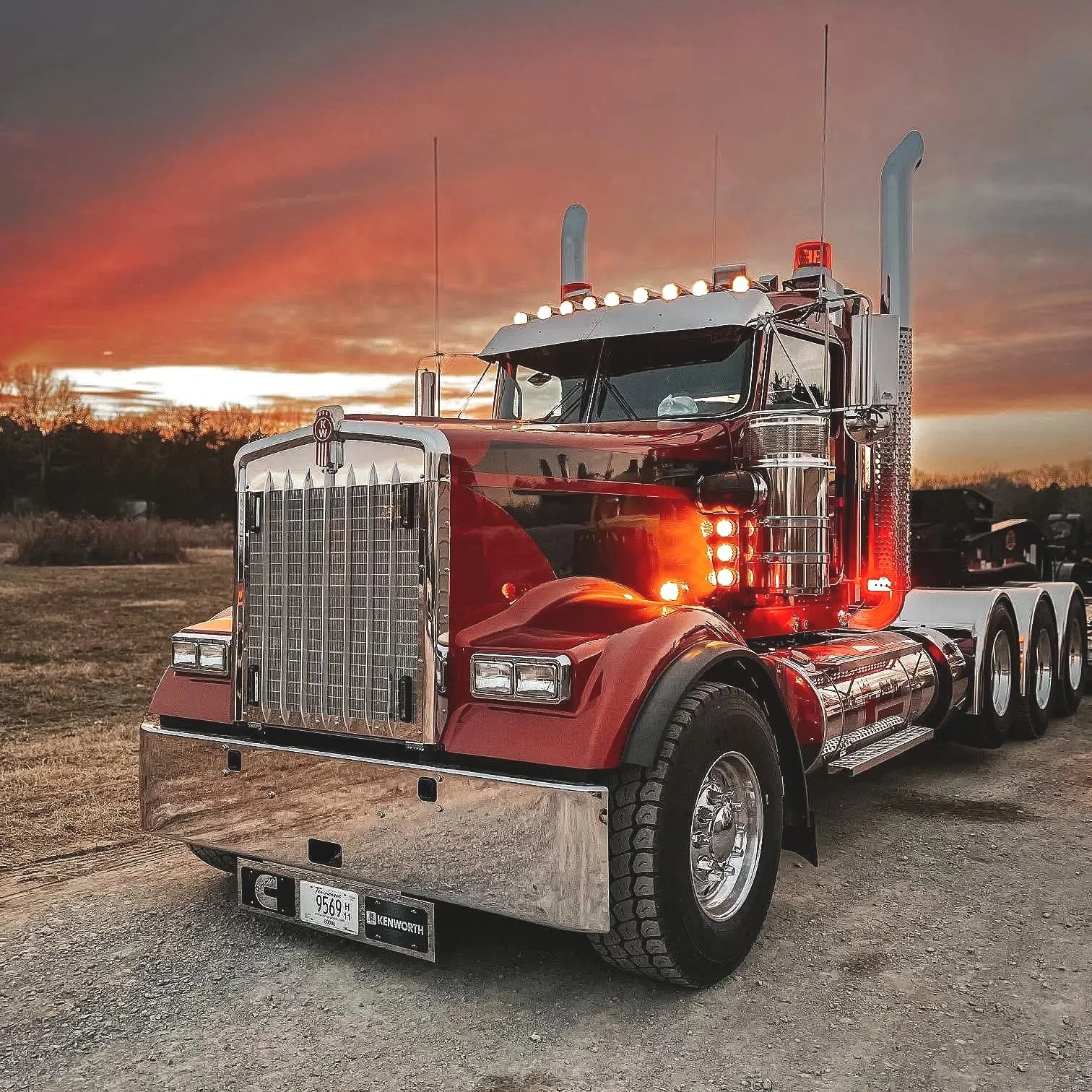 A shiny red semi-truck parked outdoors during sunset with a colorful sky, showing large front grille, multiple lights, and exhaust pipes.