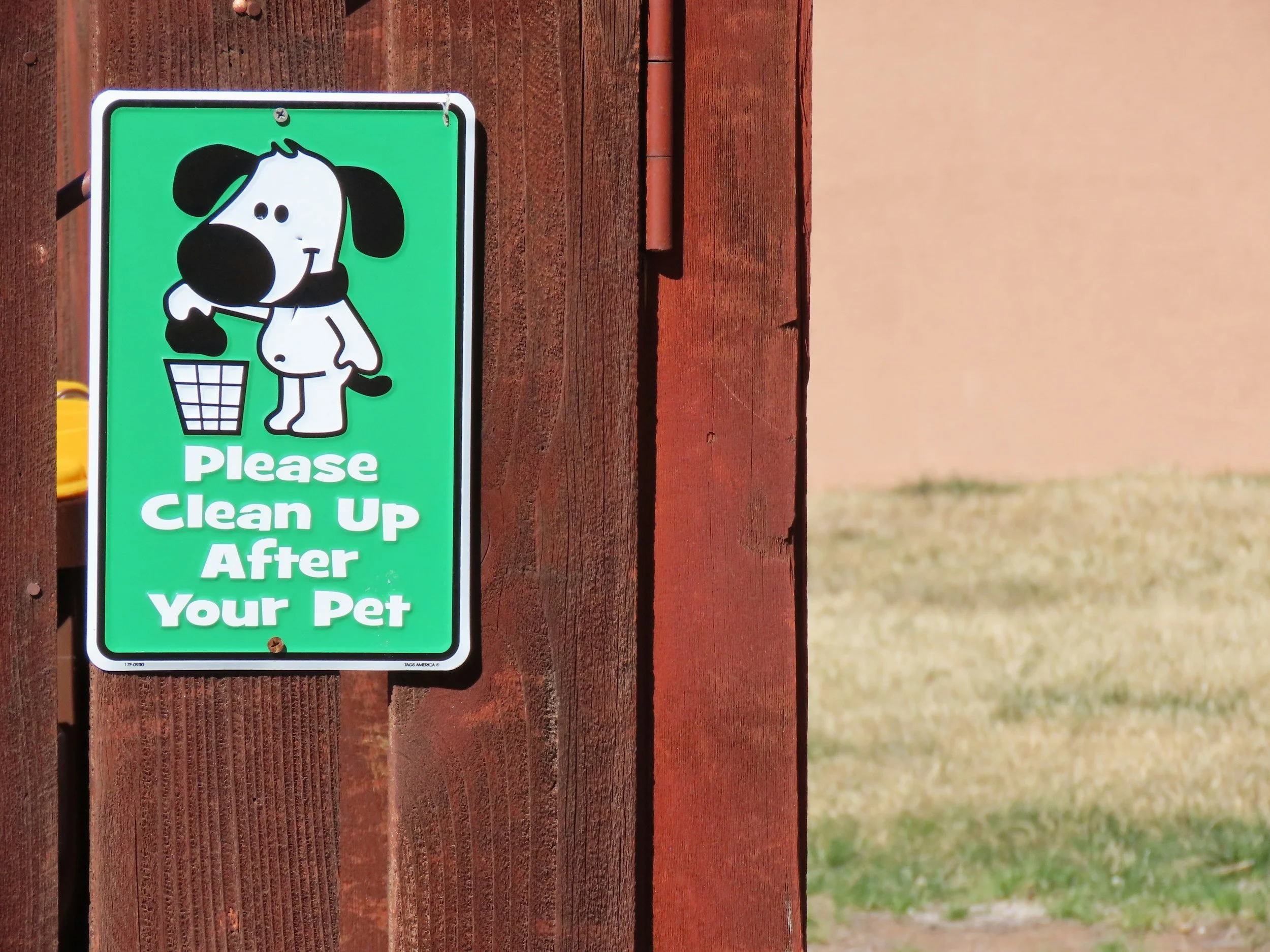 A green sign attached to a wooden fence shows a cartoon dog holding a paw in front of a trash bin, with the text "Please Clean Up After Your Pet".