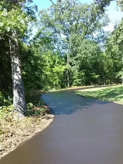 A freshly paved asphalt road winds through a green, wooded area with trees and grass on either side.