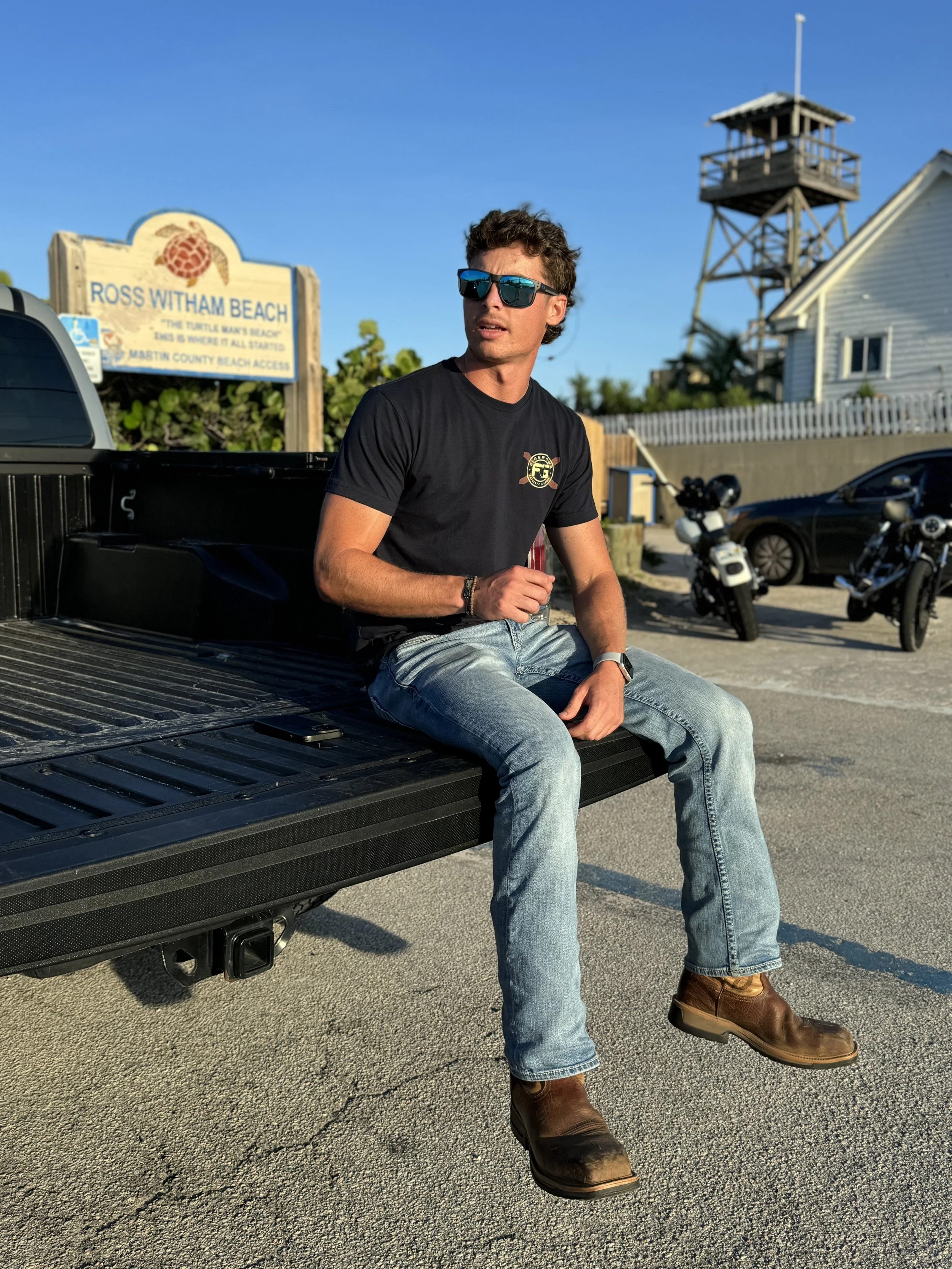 A young man with curly brown hair wearing dark sunglasses, a black t-shirt, light blue jeans, and brown boots, sitting on the edge of a black pickup truck's truck bed at Ross Witham Beach. There is a sign for Ross Witham Beach in the background along with a wooden watchtower, cars, and motorcycles parked nearby, and a bright blue sky.