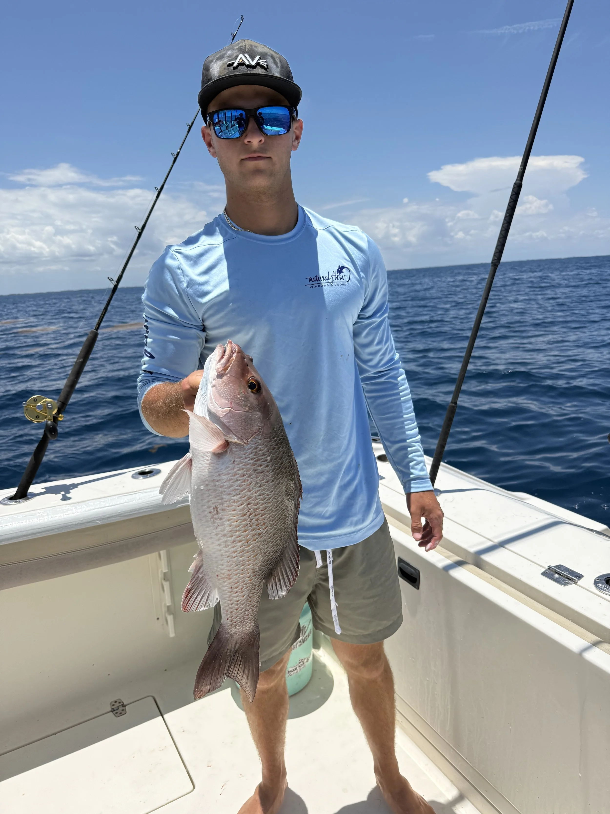 A man wearing a black cap with the word 'AVE' and reflective sunglasses holding a large fish on a boat with a clear blue sky and ocean in the background.