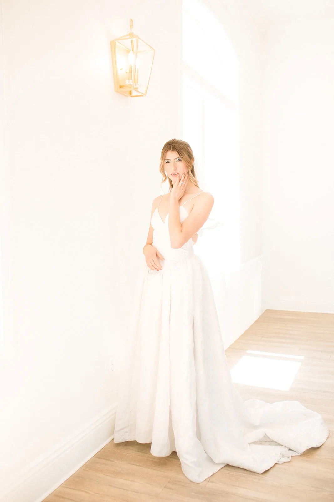 A young woman in a white dress standing in a bright room with white walls, wooden floor, and a window. She has light brown hair and is touching her face with one hand, smiling softly.
