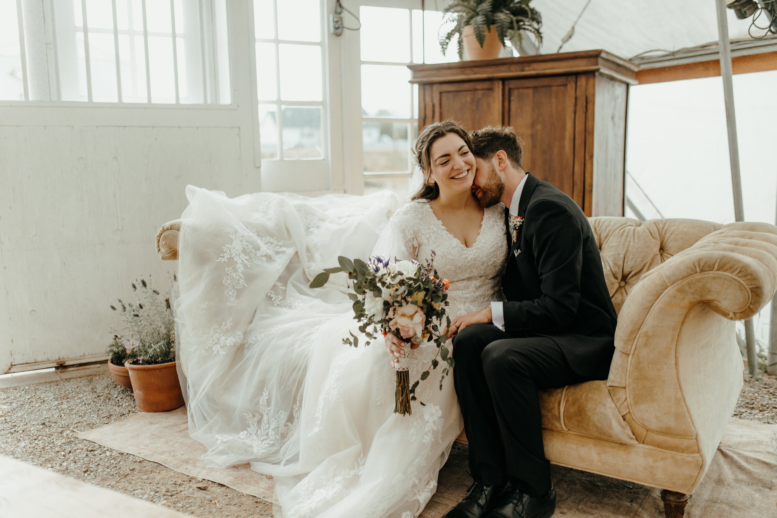 A bride and groom sitting on a vintage sofa in a rustic indoor setting, smiling and embracing, with the bride holding a bouquet of flowers.