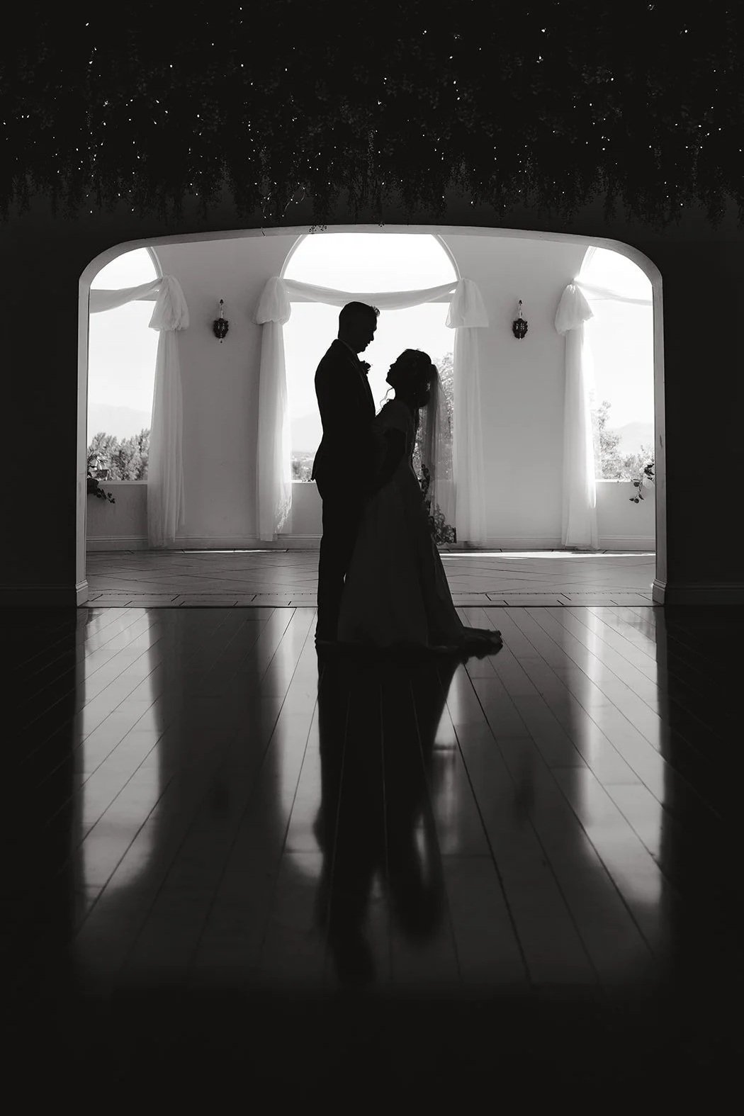 Silhouette of a couple, likely bride and groom, standing close together in a wedding venue with large windows, with reflections on the polished wooden floor.