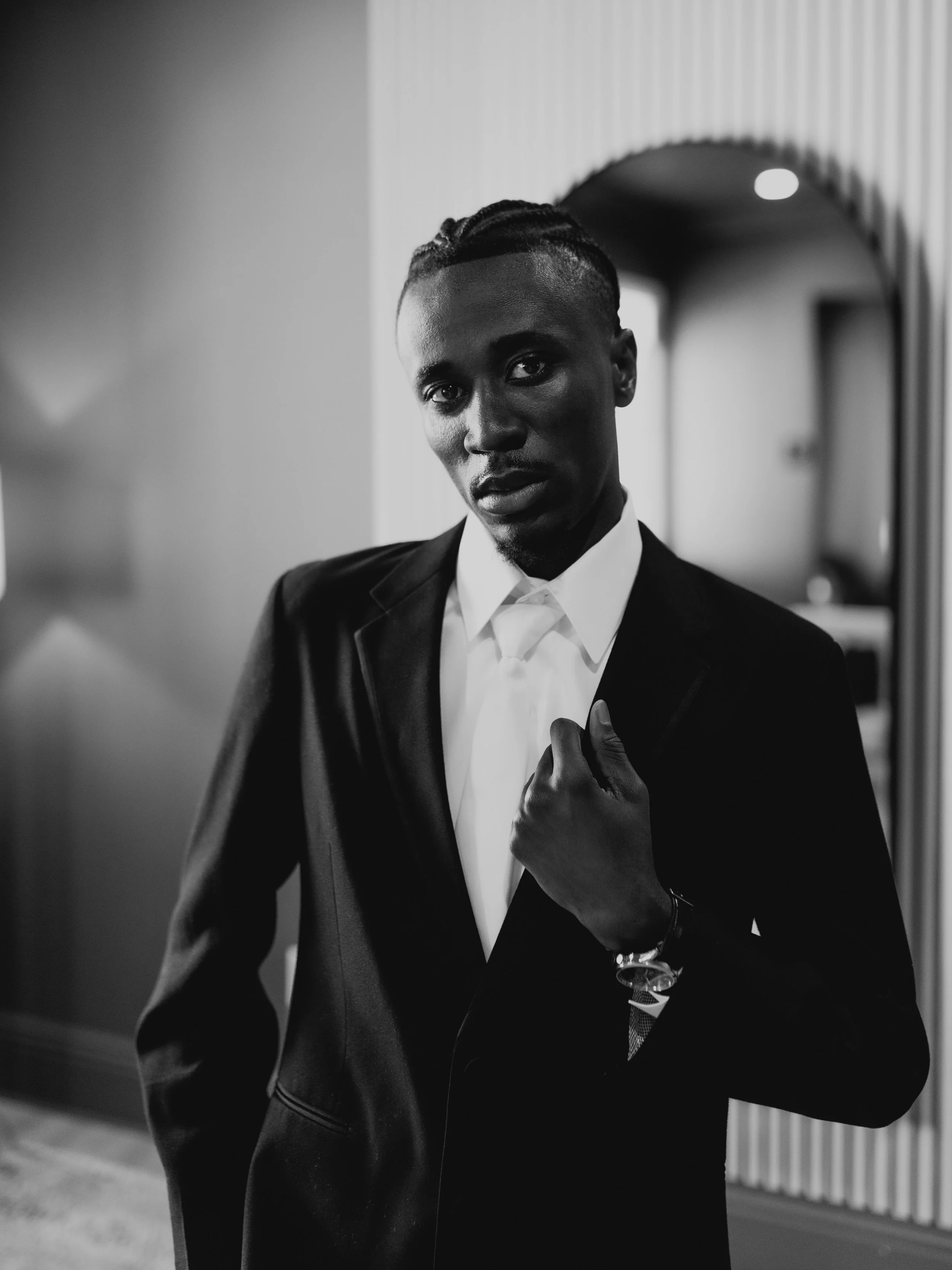 A black and white photo of a man with braided hair, wearing a suit and tie, adjusting his sleeve in an indoor setting with a blurred background.