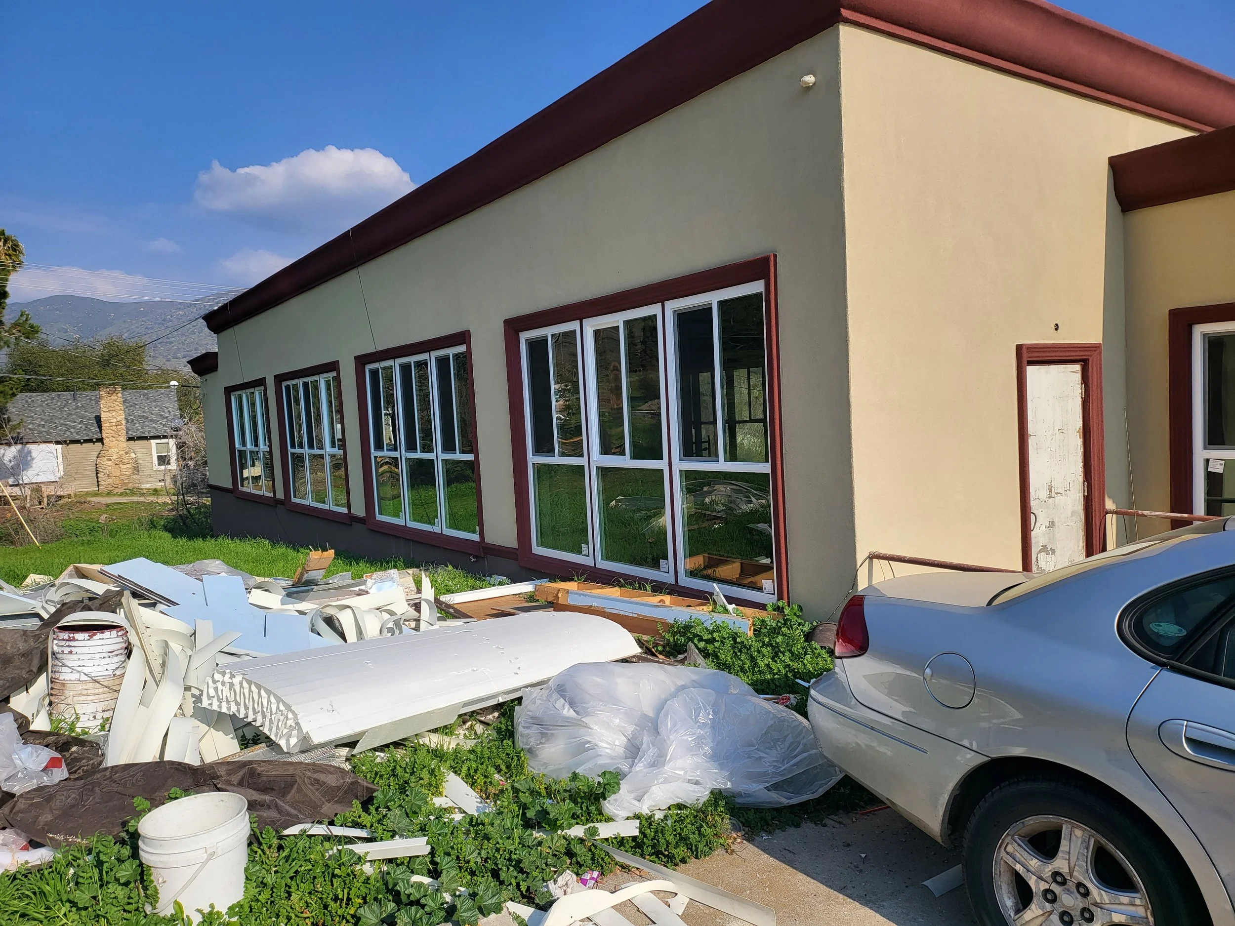 A house with large windows and a yellow exterior, surrounded by construction debris, including white panels, plastic sheets, and buckets, with two silver cars parked in the driveway. In the background, there are neighboring houses and mountains under