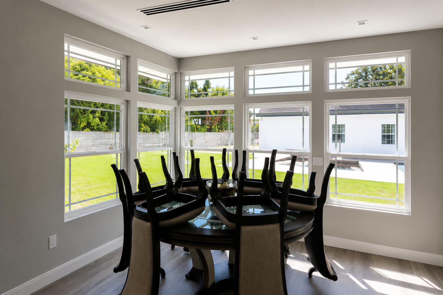 A dining room with a round table surrounded by chairs, situated near large windows that let in natural sunlight and offer a view of a green backyard.