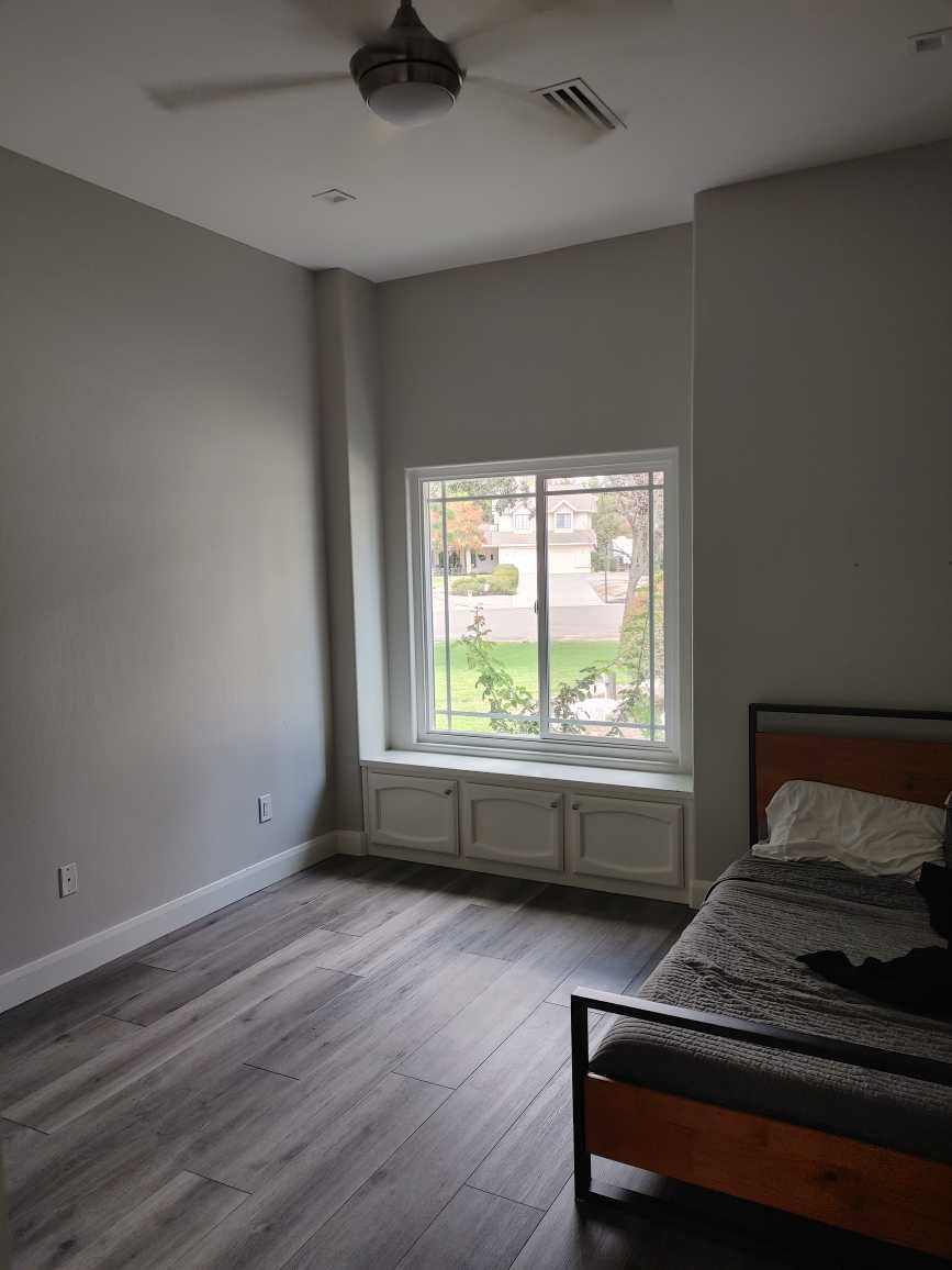 A minimally furnished bedroom with a large window, gray walls, wood flooring, and a bed with a gray bedspread and black clothing on it.