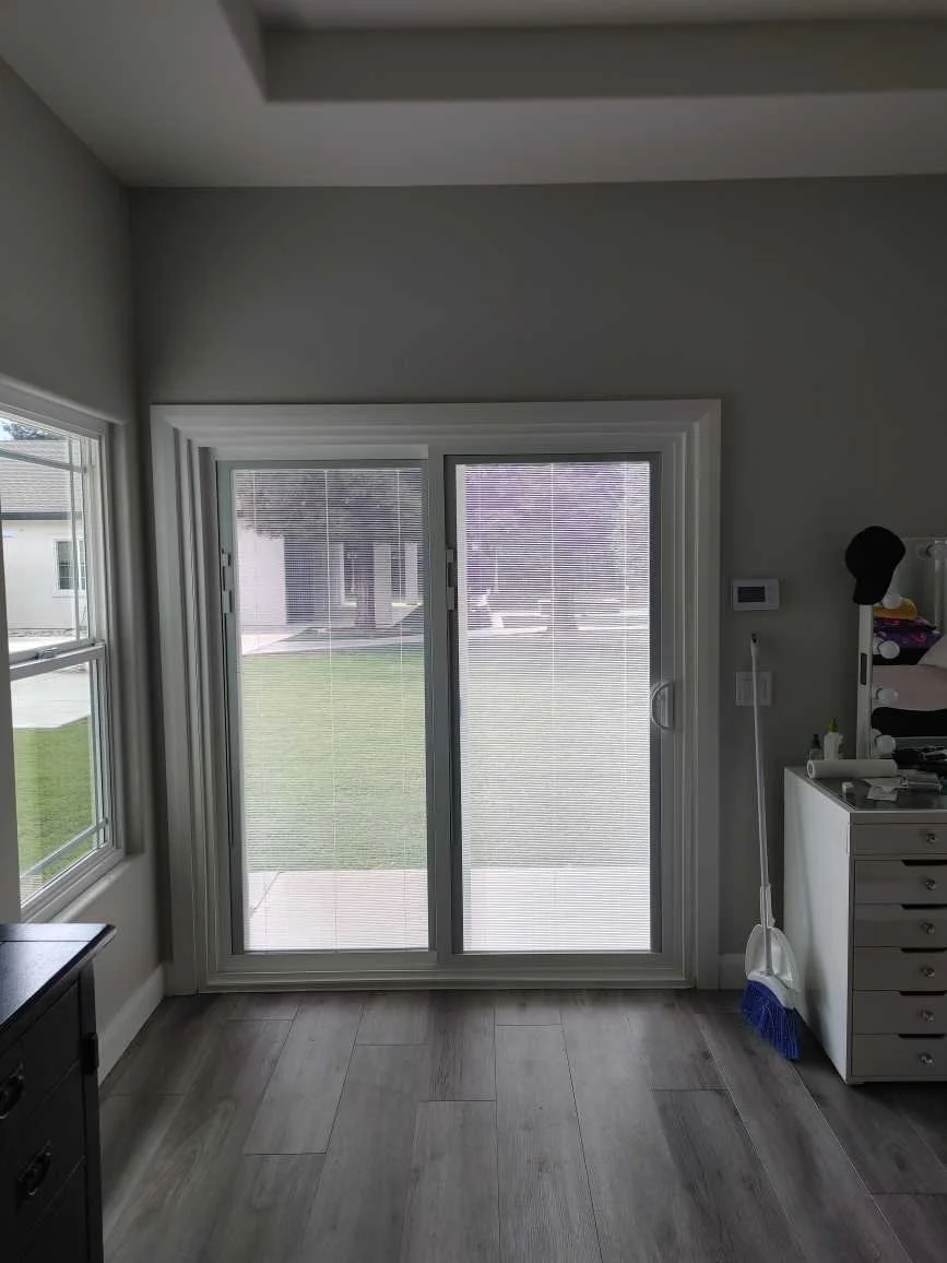Interior view of a room with sliding glass door with blinds, looking out to a lawn and neighboring houses, with a white chest of drawers, a broom, and various items on top.