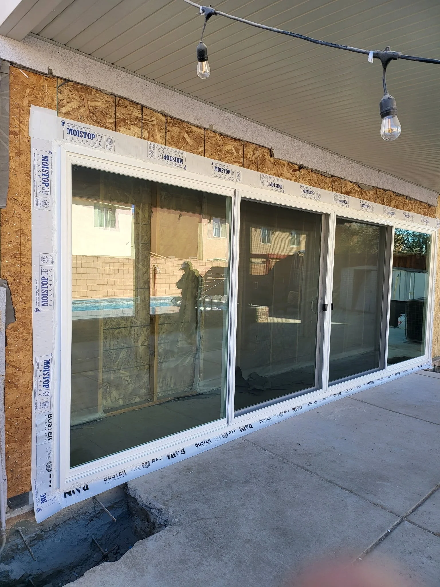 Construction of a house with sliding glass doors, with building materials and tools present, and covered outdoor space with string lights.