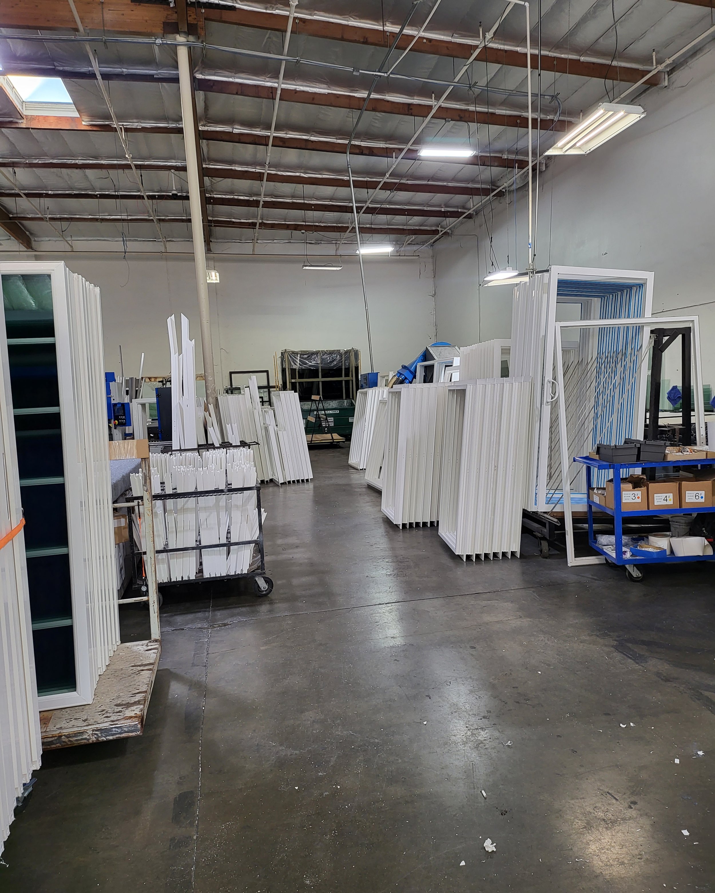 Empty industrial warehouse with metal framing and multiple windows, with various metal and plastic items arranged in rows along the sides and center.