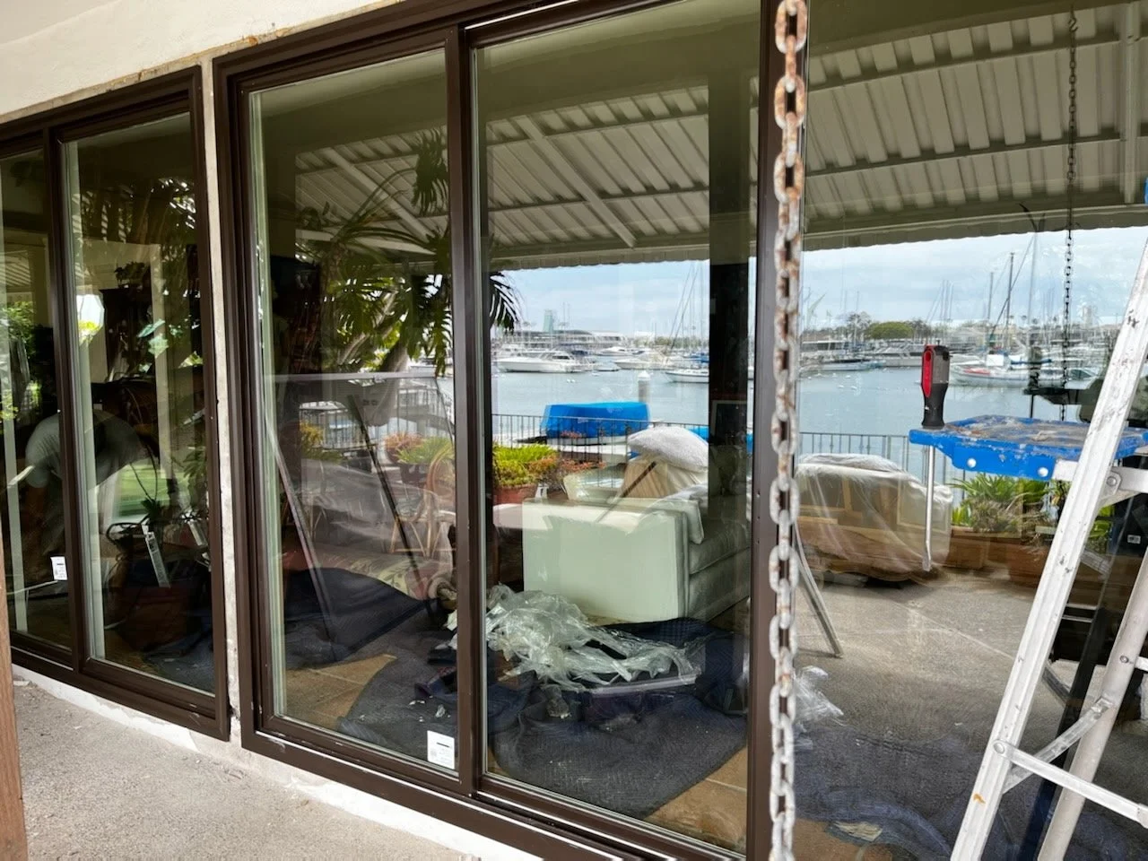 View through large glass sliding doors overlooking a marina with boats, and a covered outdoor patio with furniture, plants, a ladder, and construction tools.