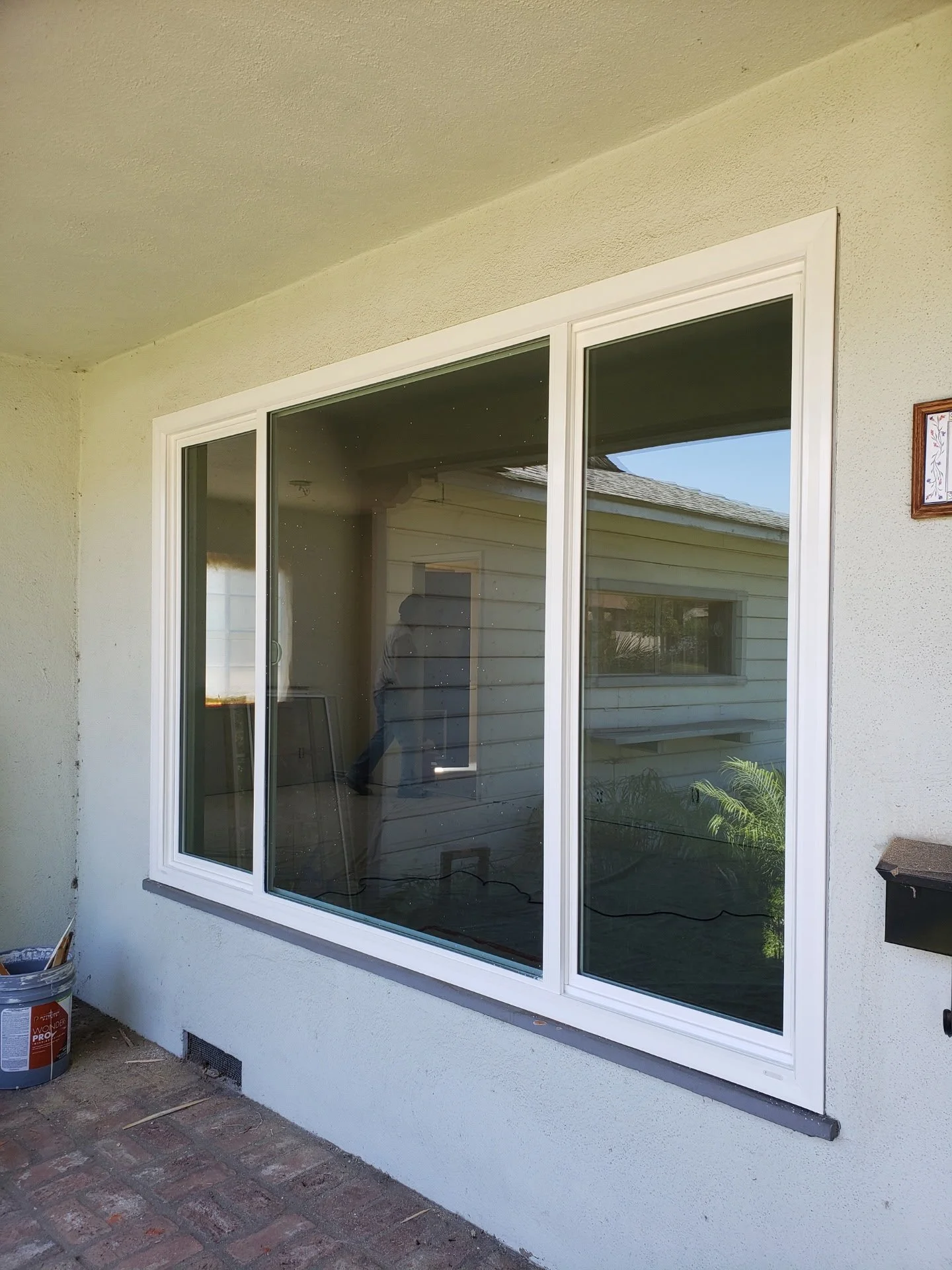 A large white-framed sliding glass door on a white stucco house wall, with a reflection of the interior room and a person taking the photo. There is a small plant visible in the reflection and a welcome sign on the wall to the right.