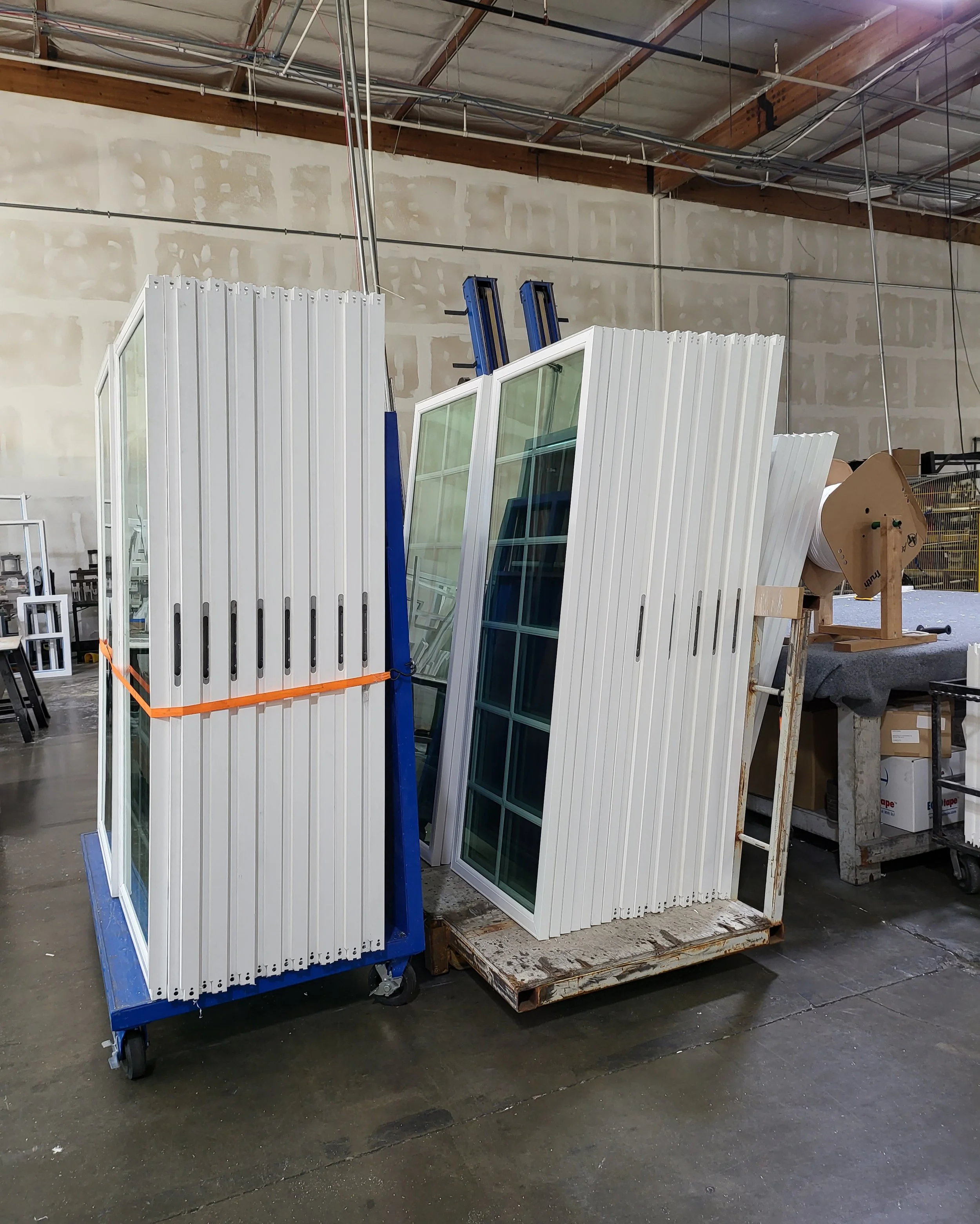 Stacks of white window frames with glass panes leaning against a blue rolling cart inside a warehouse or workshop with unfinished walls and a metal roof.
