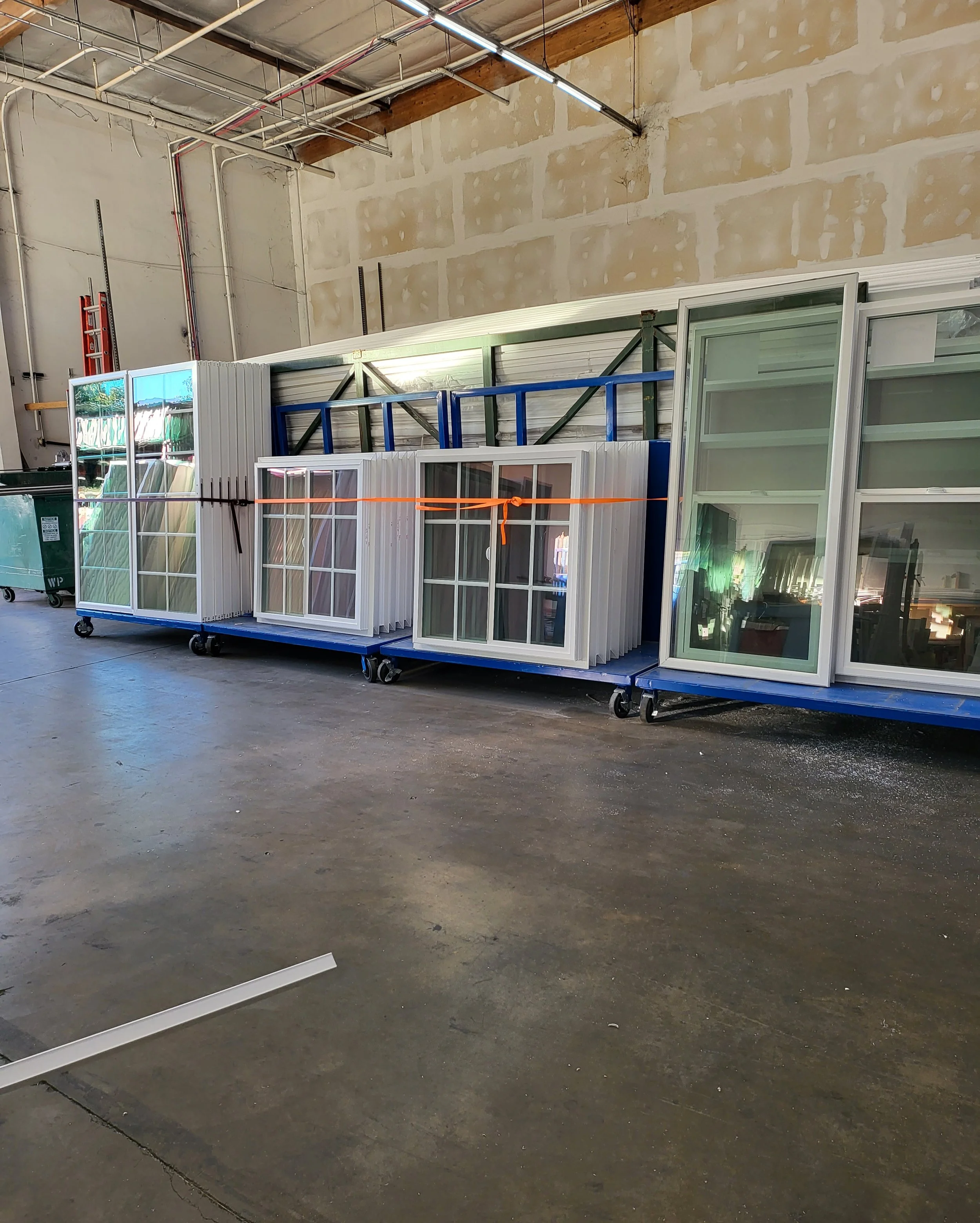 Indoor warehouse with unfinished walls, ceiling pipes, and various windows and panels stacked on blue carts, some tied with orange straps, on a concrete floor.