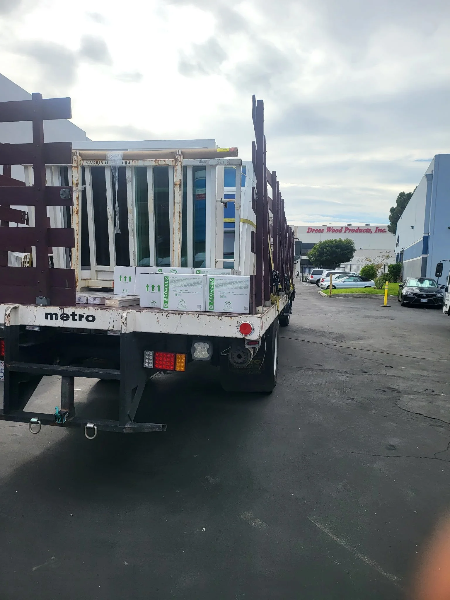 A flatbed truck parked in a parking lot with building and trees in the background. The truck carries boxes of windows or glass panels.
