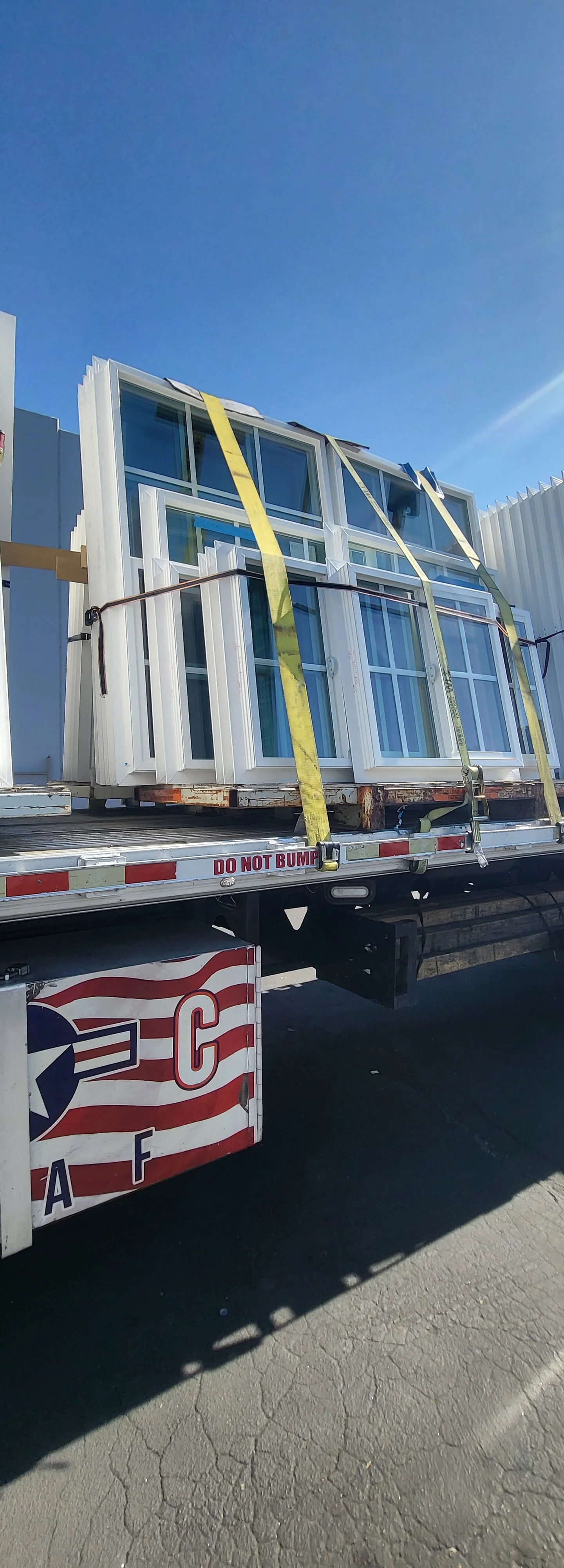 Delivery truck carrying large white windows strapped down, with a blue sky in the background.
