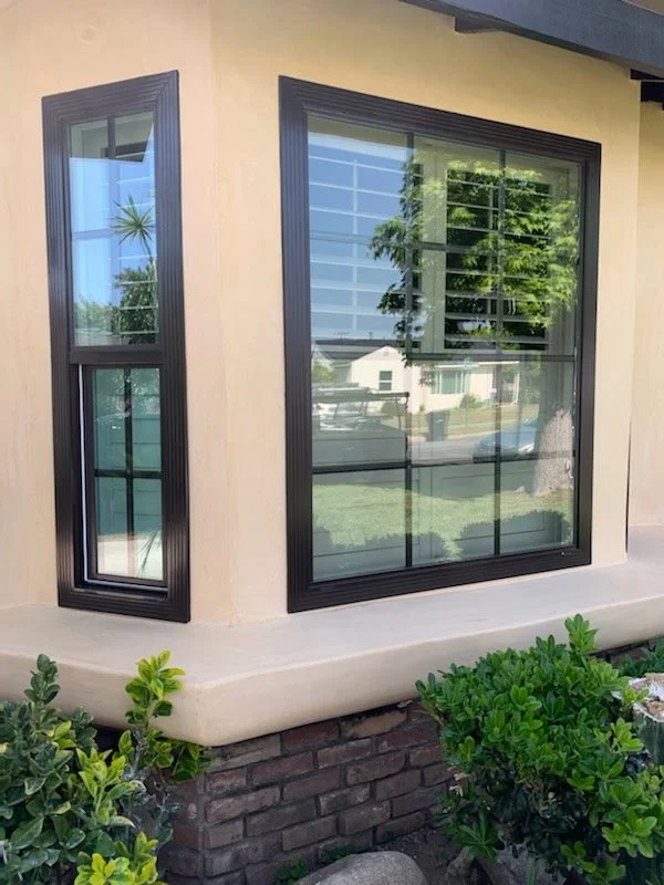 Close-up of a modern house exterior featuring a large, rectangular window with black trim and a smaller, vertical window also with black trim. The house's wall is light beige, with a brick foundation and green shrubs in the foreground. The window ref