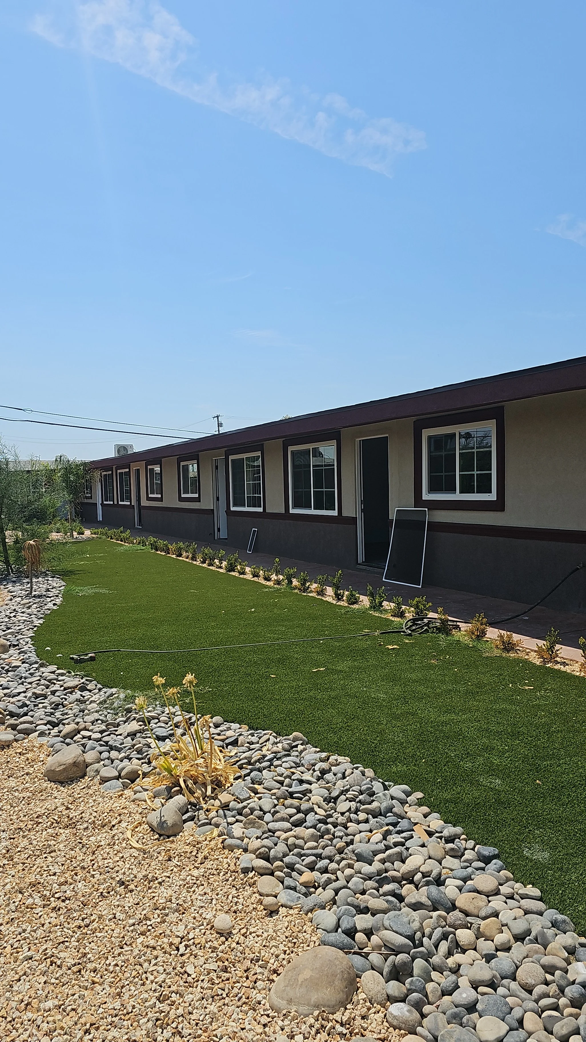 A row of beige and brown single-story houses with multiple windows and doors, set in a landscaped yard with green grass, small plants, and decorative rocks. The sky is blue with wispy clouds.