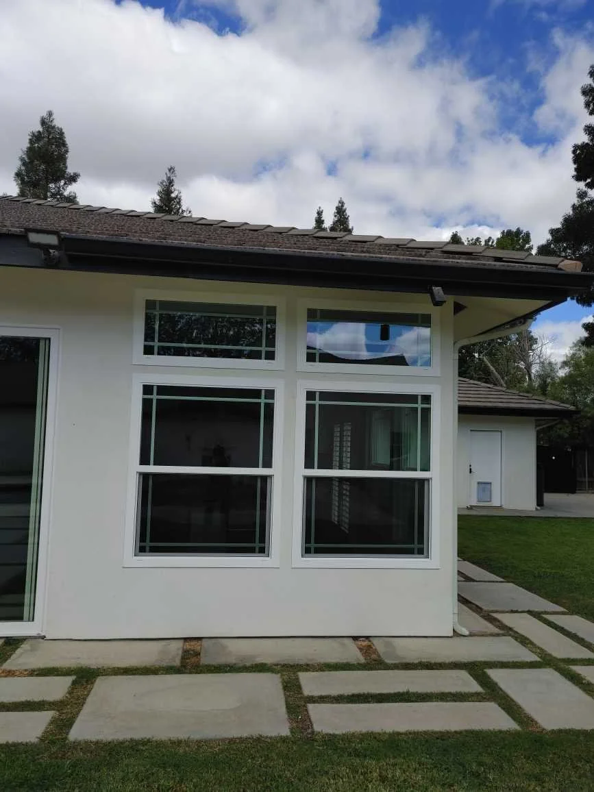 A house with white exterior walls, multiple large rectangular windows, a stone pathway on the lawn, and a partly cloudy sky above.
