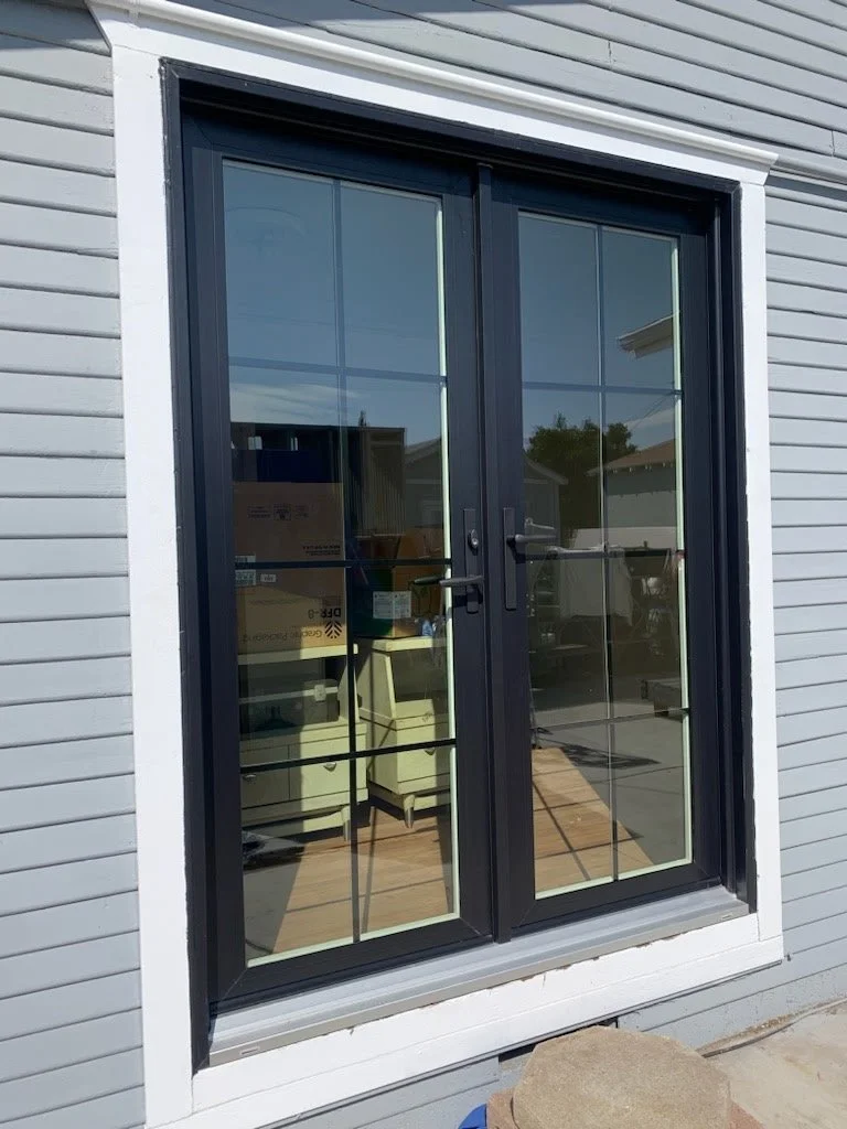 Double glass doors with black frames and handles, reflecting surrounding houses and sky, installed on a house with light gray siding and white trim.