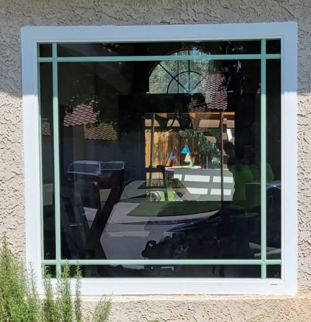 Reflected view of a backyard with a playground set, patio, trees, and a wooden fence, seen through a large window in a beige stucco house.
