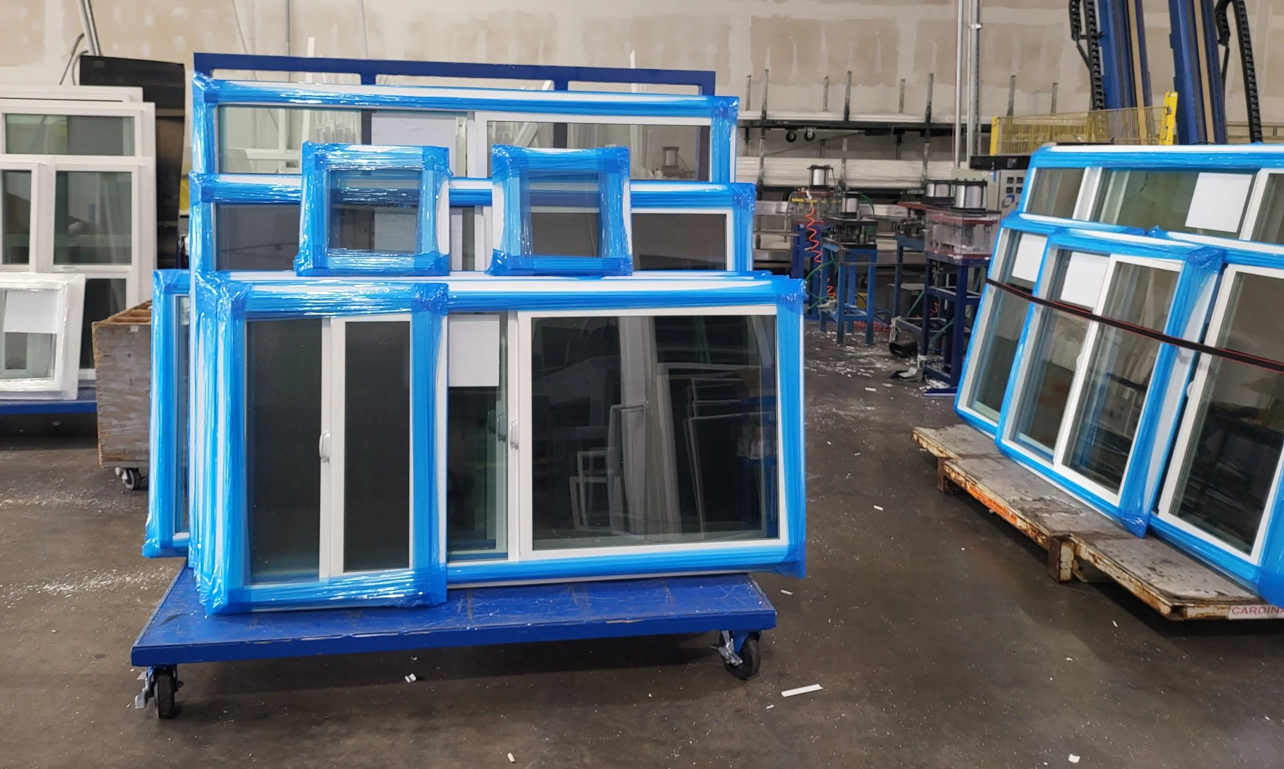Stacks of new white-framed windows with blue plastic wrapping in an industrial workshop.