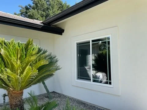A white house with a window, green palm plant, and gravel ground outside.