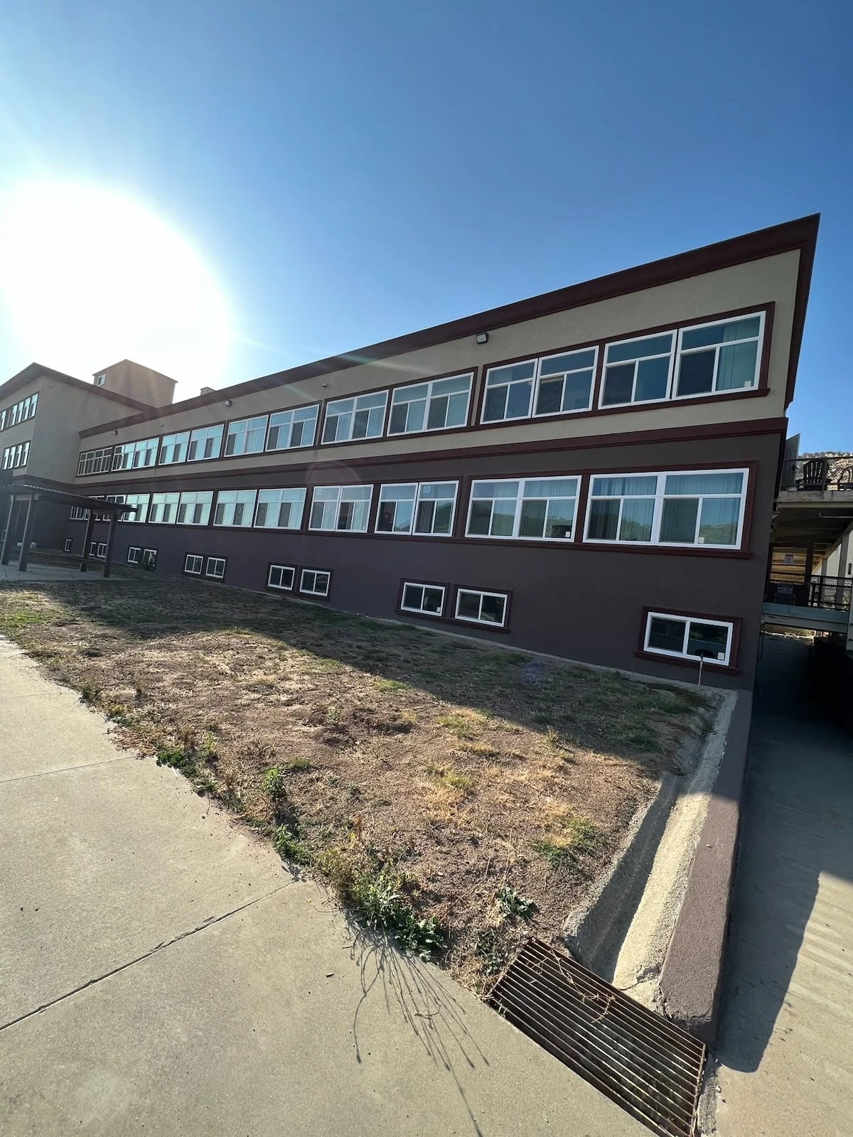 Modern multi-story building with large windows and an angled roof under a clear blue sky, with a patch of dirt and concrete sidewalk in foreground.