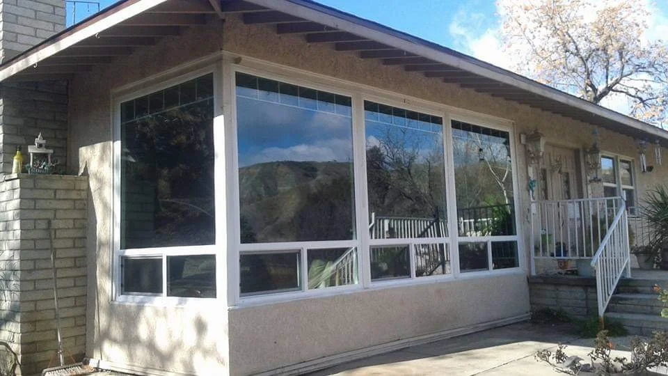 A house with a large enclosed porch or sunroom featuring big glass windows, a brick exterior on one side, and stairs leading up to the door. There are trees and hills reflected in the windows, and a small garden area in front.