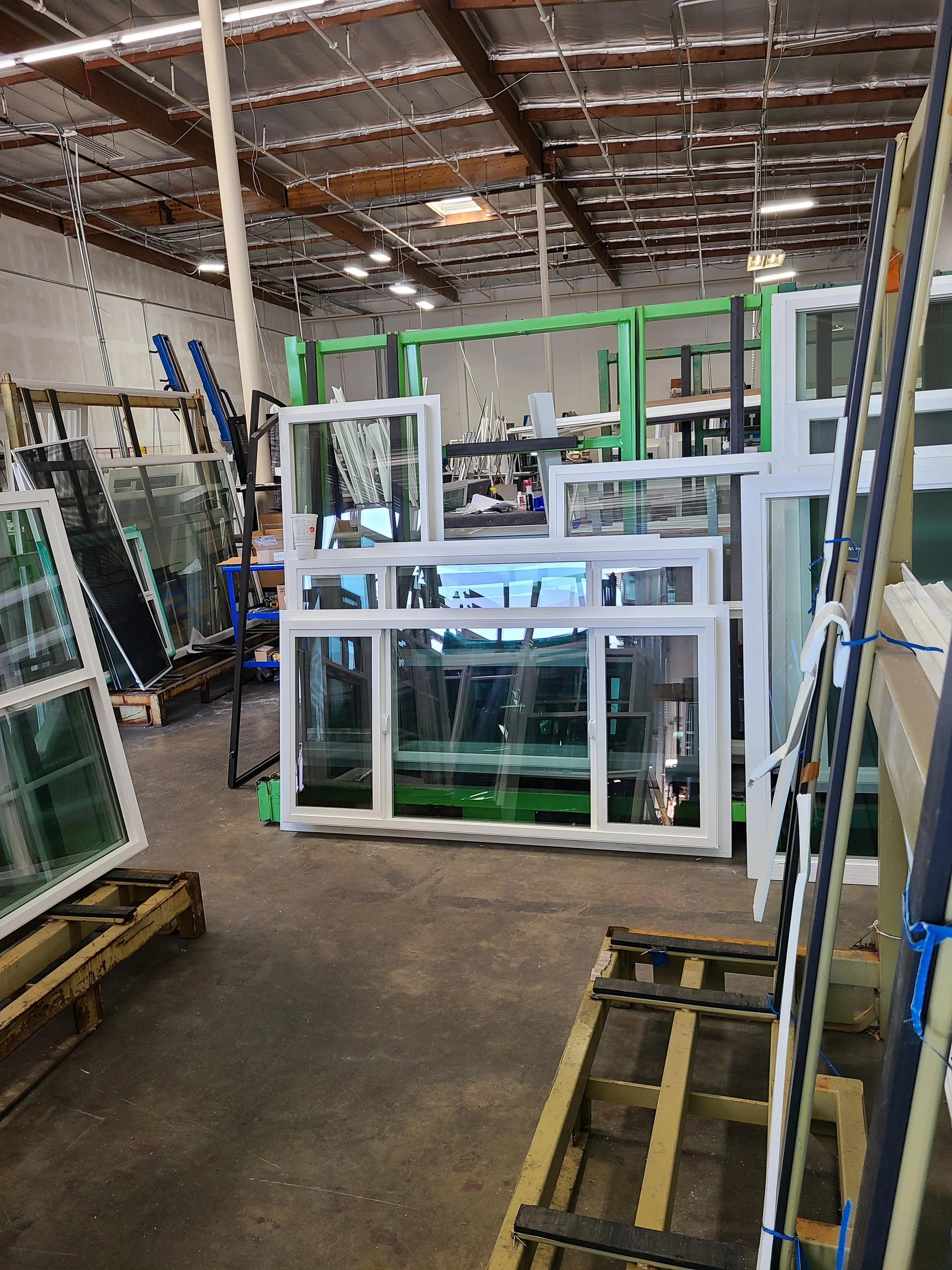 A storage area filled with various white window frames, some leaning against each other and the wall, with metal shelves and construction supplies scattered on the floor inside a warehouse with unfinished ceiling and exposed beams.