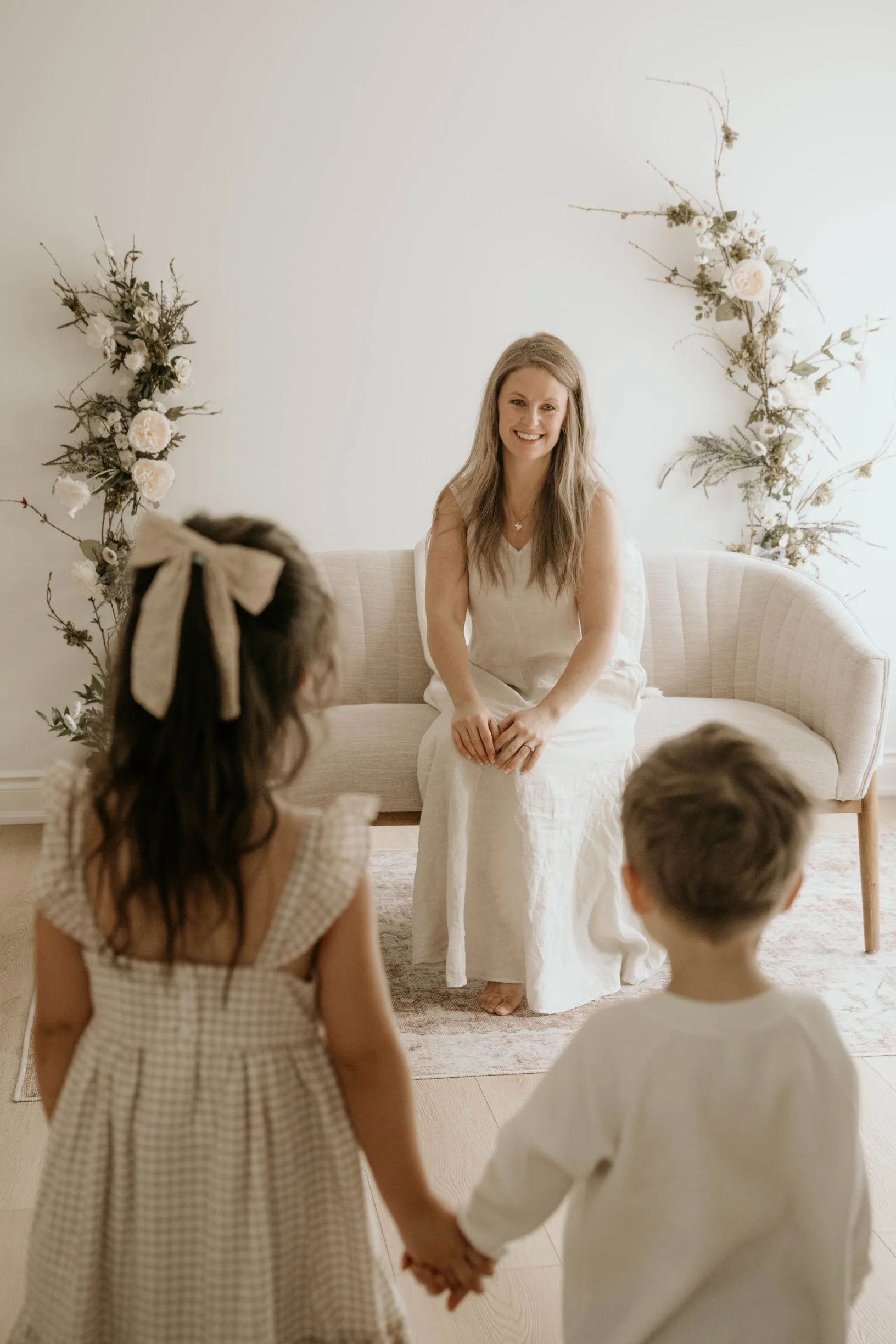 A young woman with long blonde hair, wearing a white dress, sitting on a cream-colored couch, smiling at children in front of her. The scene features a minimalist, floral-decorated background with white and green foliage. Two children, a girl with a beige bow in her hair and a boy, are holding hands and facing the woman.