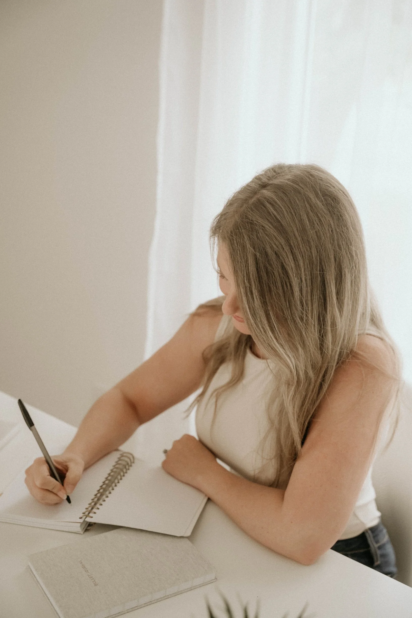 A young woman with long, wavy blonde hair writing in a spiral notebook at a white table, with a book titled 'PERFECTLY IMPERFECT' on the table.