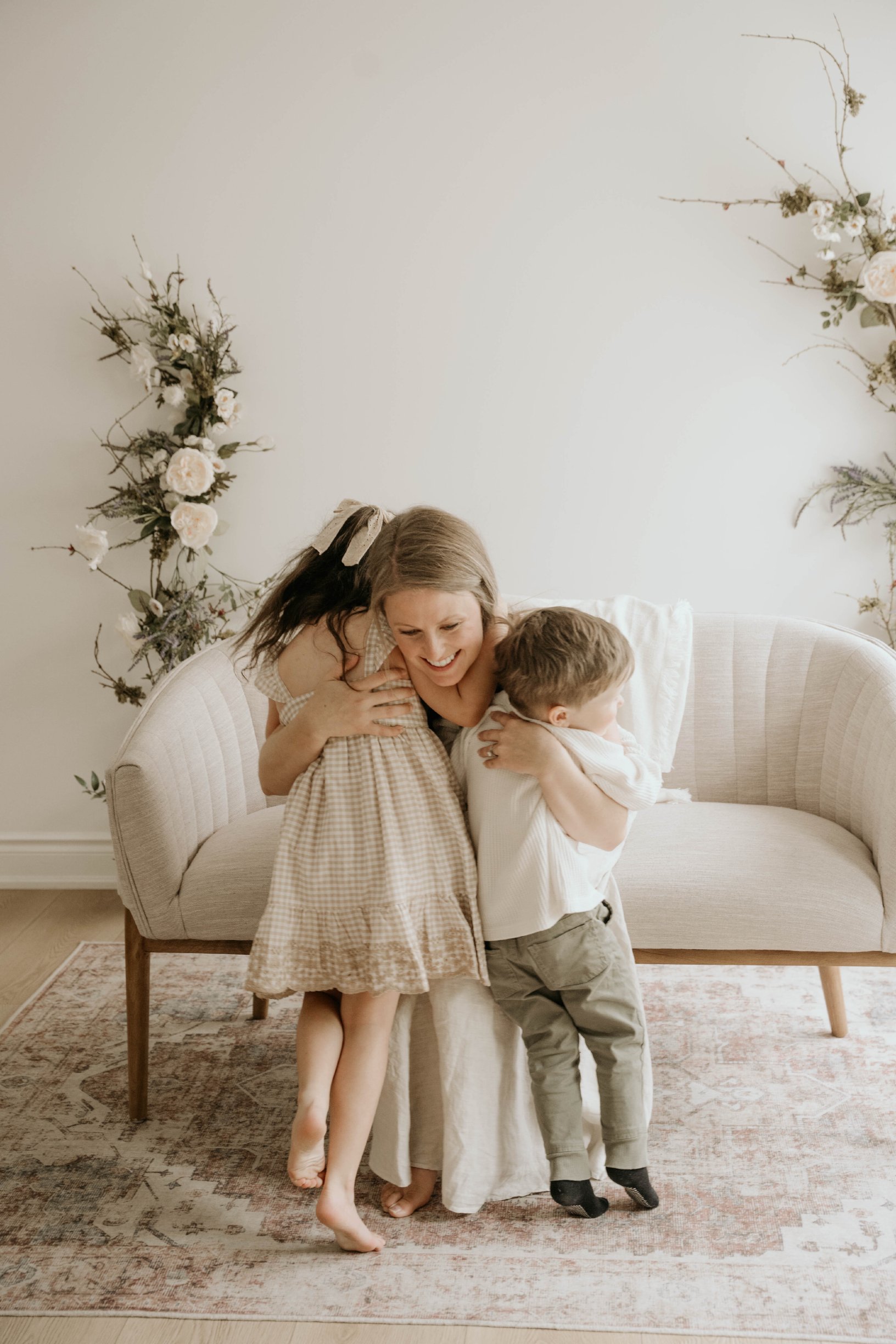 A woman with short blonde hair, wearing a white dress, is sitting on a light-colored couch and embracing two young children. One child, a girl with long hair tied with a bow, is standing on her lap, and the other, a boy with short hair, is standing next to her with one arm around her neck. They are all smiling and sharing a joyful moment in a cozy, decorated living room with floral wall accents and a patterned rug.