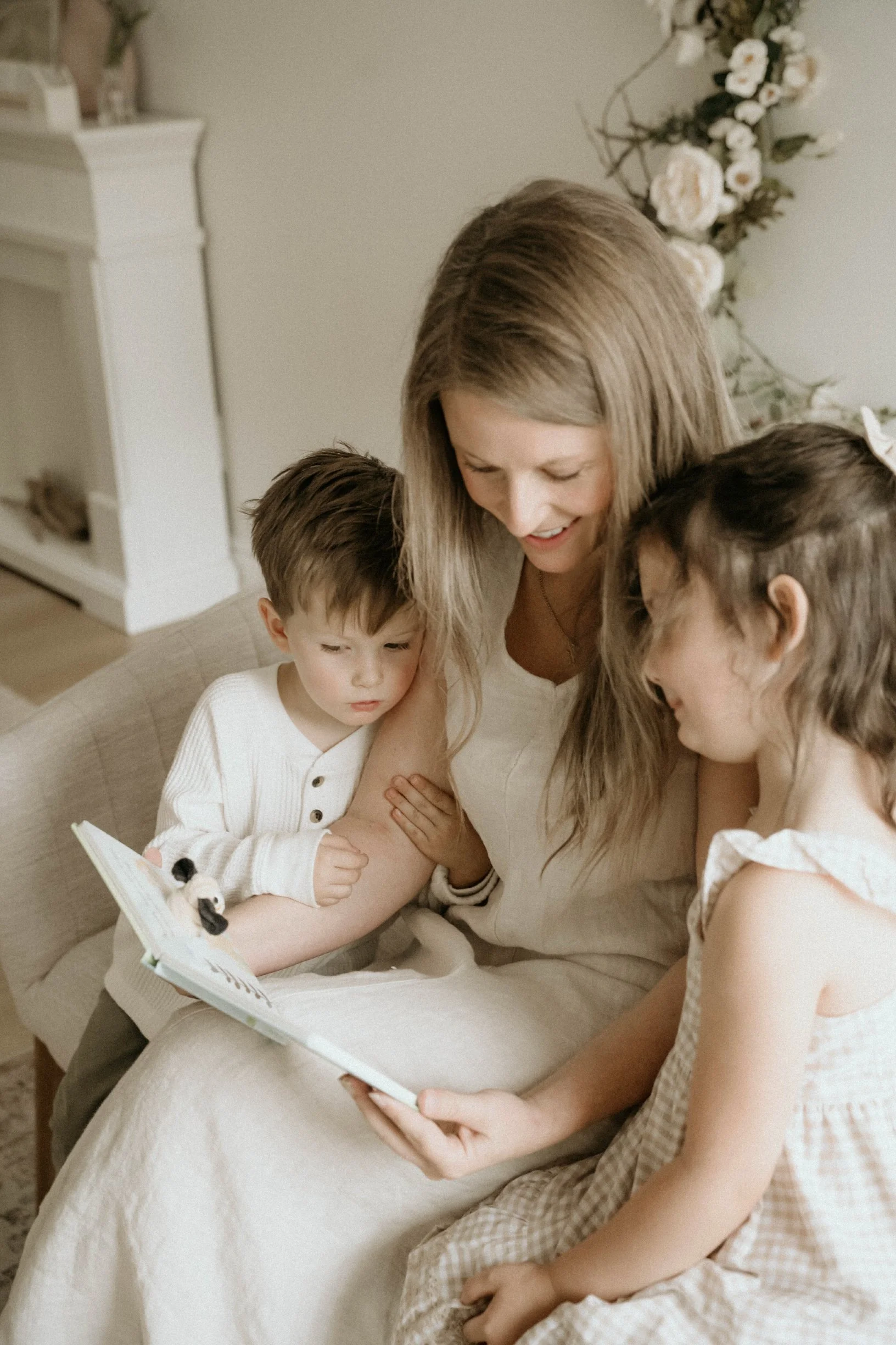 A woman with long blonde hair sitting on a beige couch, reading a children's book to two young girls. One girl has short brown hair, and the other has long brown hair with a bow. The background features a white fireplace mantel and a floral arrangement with white flowers.