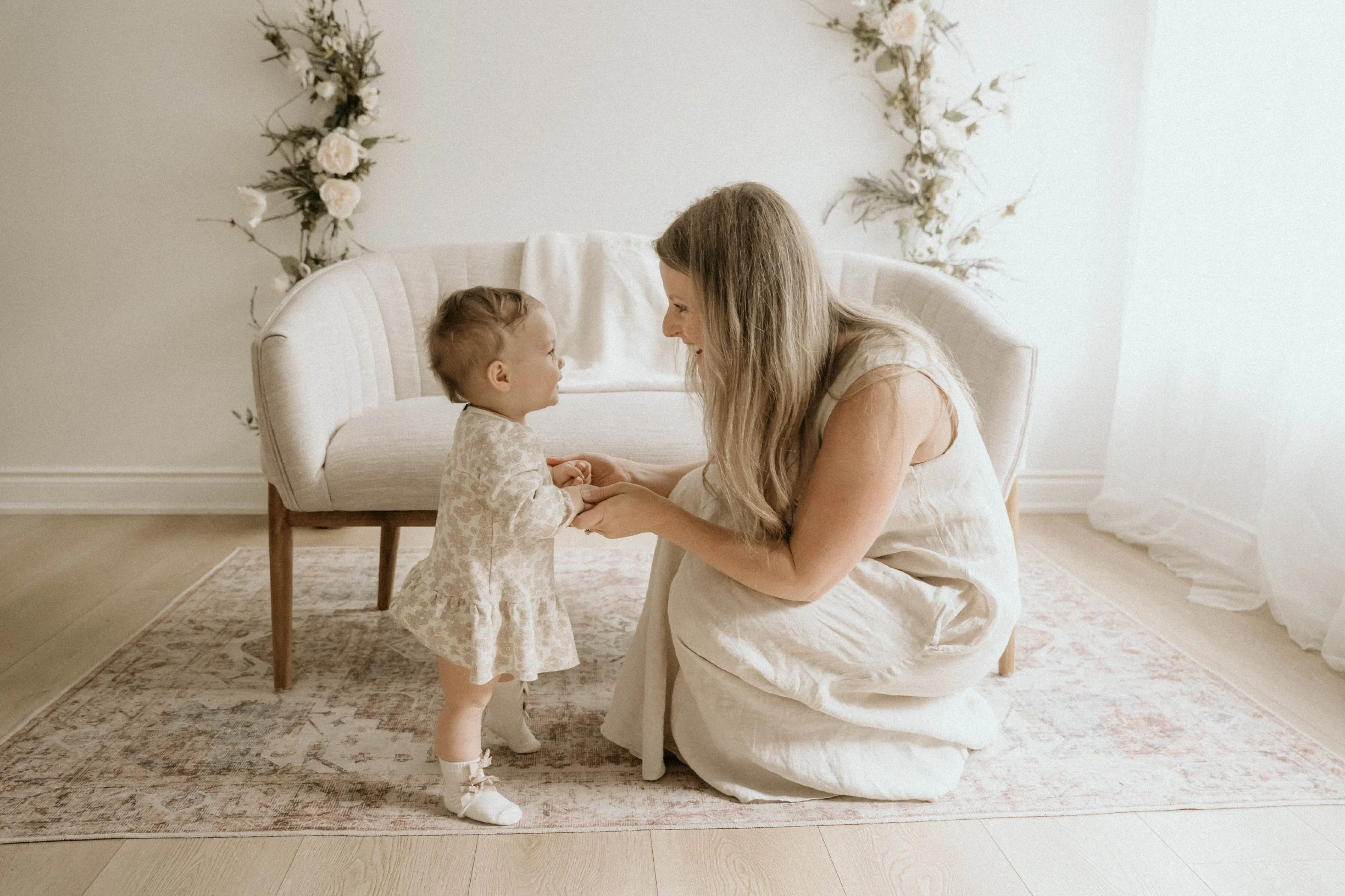 A woman and a young girl smiling and holding hands in a living room with floral wall decorations and a beige armchair.