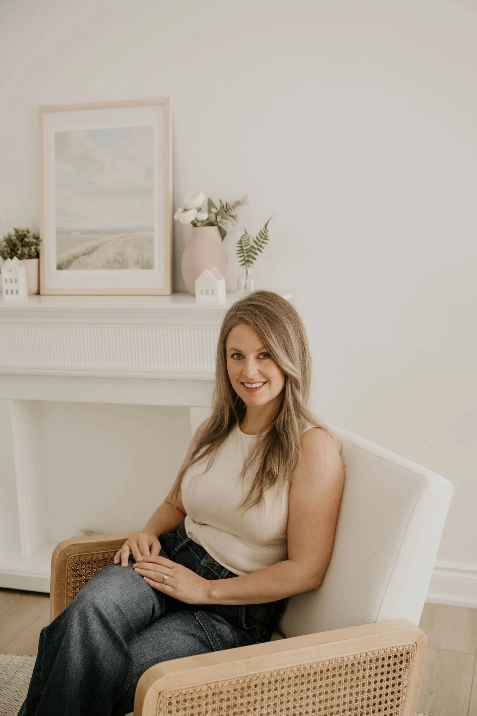 A smiling woman with long, wavy brown hair sitting on a cream-colored armchair in a cozy, well-decorated room. Behind her is a white fireplace mantel with a framed landscape painting, a pink vase with white flowers, and small decorative white houses. The room has neutral tones and a minimalist aesthetic.