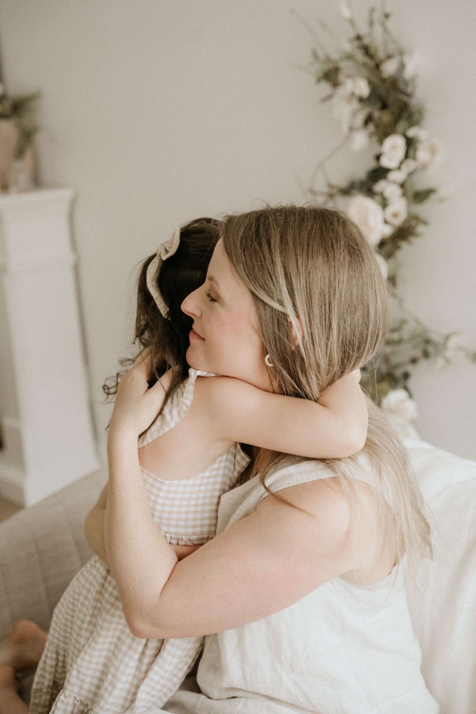 A woman and a young girl embracing and hugging each other with closed eyes. The woman has long blonde hair and is wearing a white top, the girl has dark hair with a beige bow and is wearing a beige gingham dress. They are in a room with neutral tones, and there are flowers in the background.