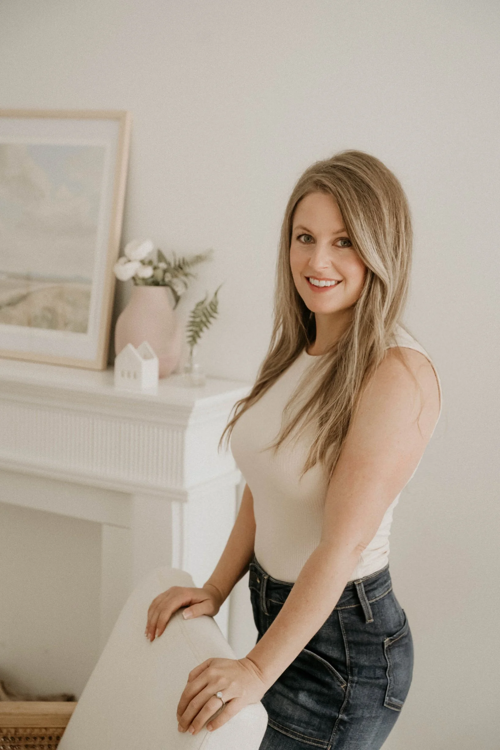 A woman with long wavy blonde hair smiling while standing next to a white chair in a room with minimal decor, including a shelf with a framed picture, a pink vase with white flowers, and a small white house ornament.