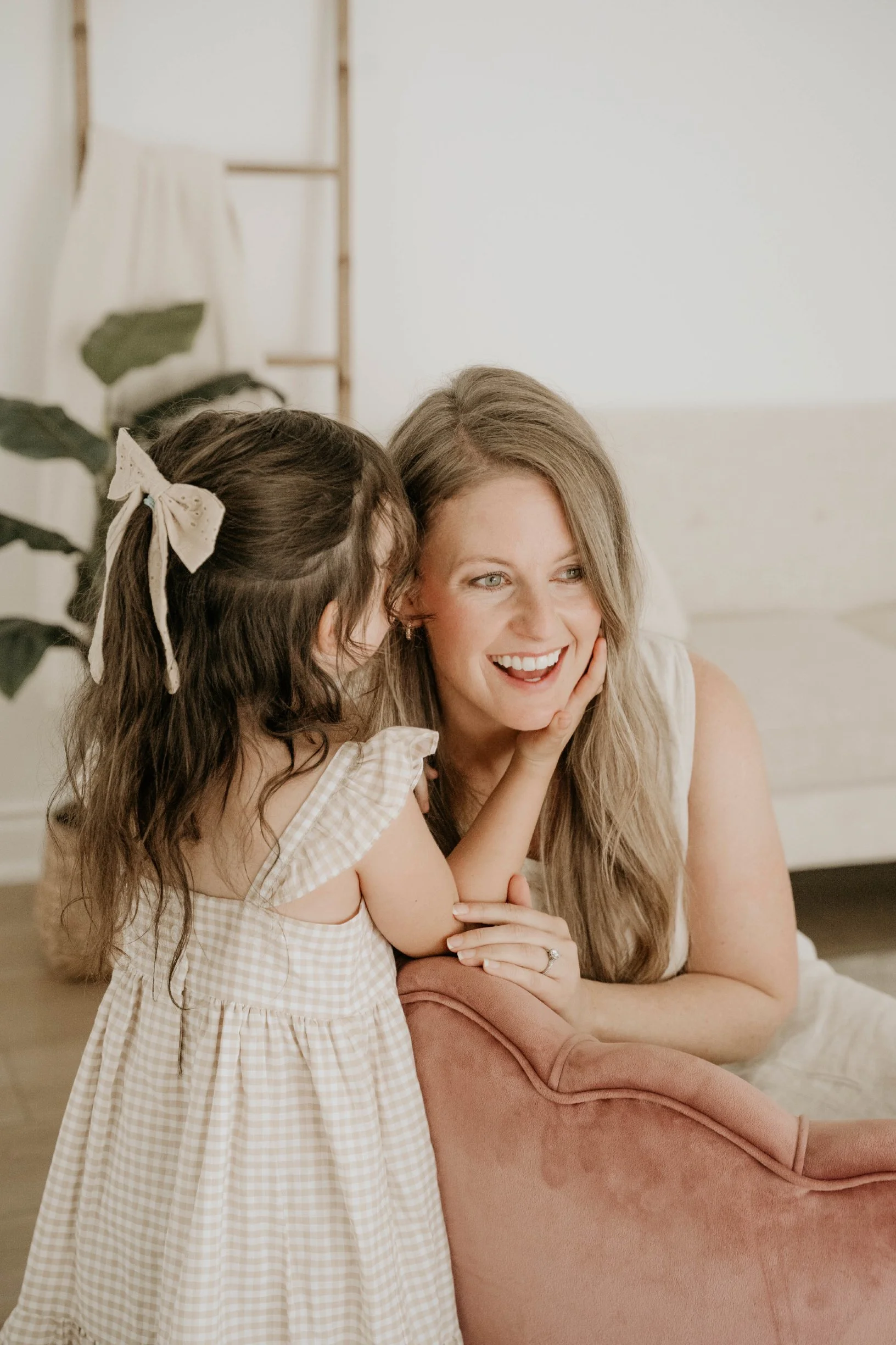 A woman with long blond hair sitting on a pink chair, smiling, as she interacts with a young girl with long dark hair tied with a cream-colored ribbon, wearing a yellow checkered dress, in a room with a white wall and a plant in the background.