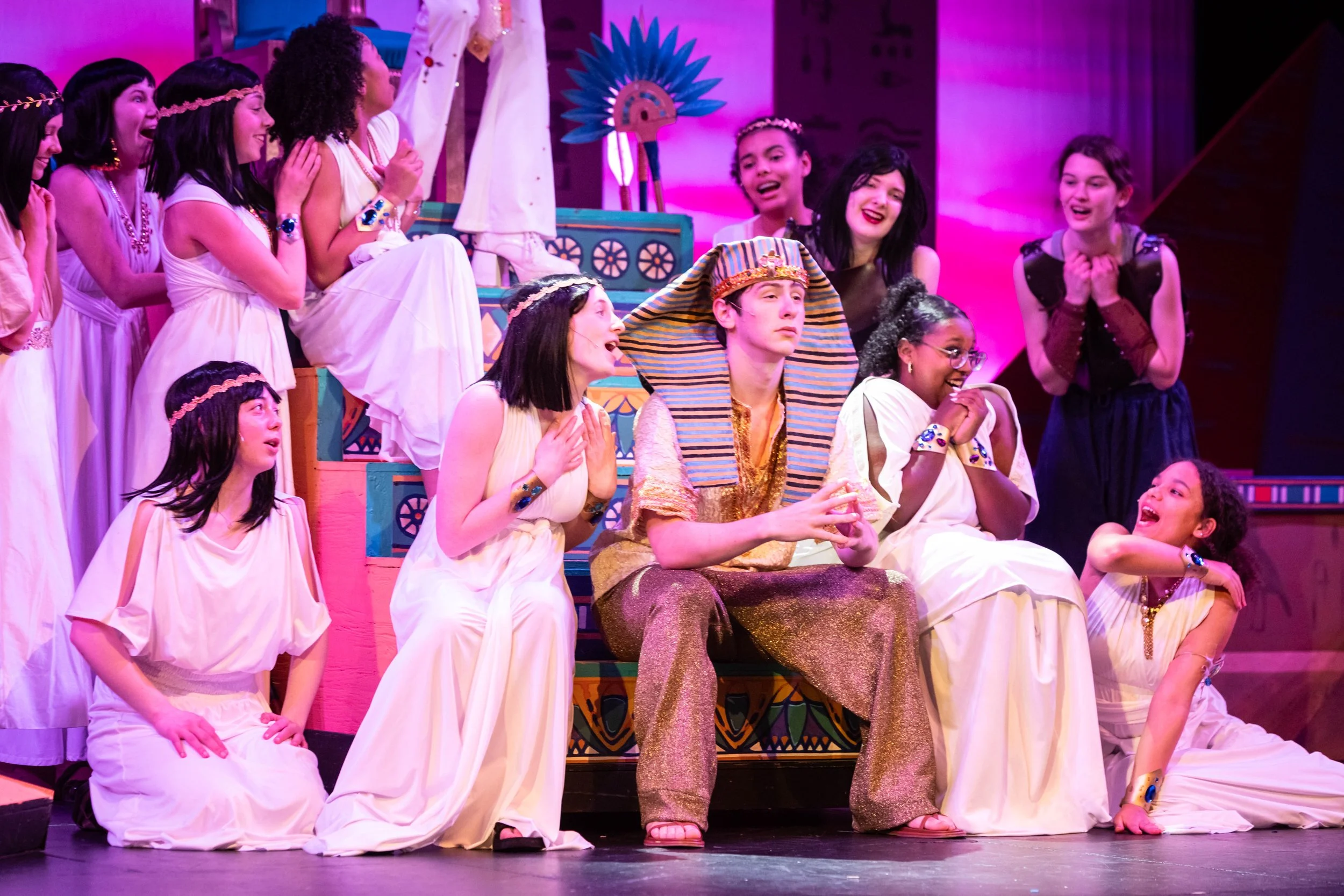 A theatrical stage with a large cast of actors dressed in ancient Egyptian costumes, some sitting and some standing, with colorful decorations and purple stage lighting.