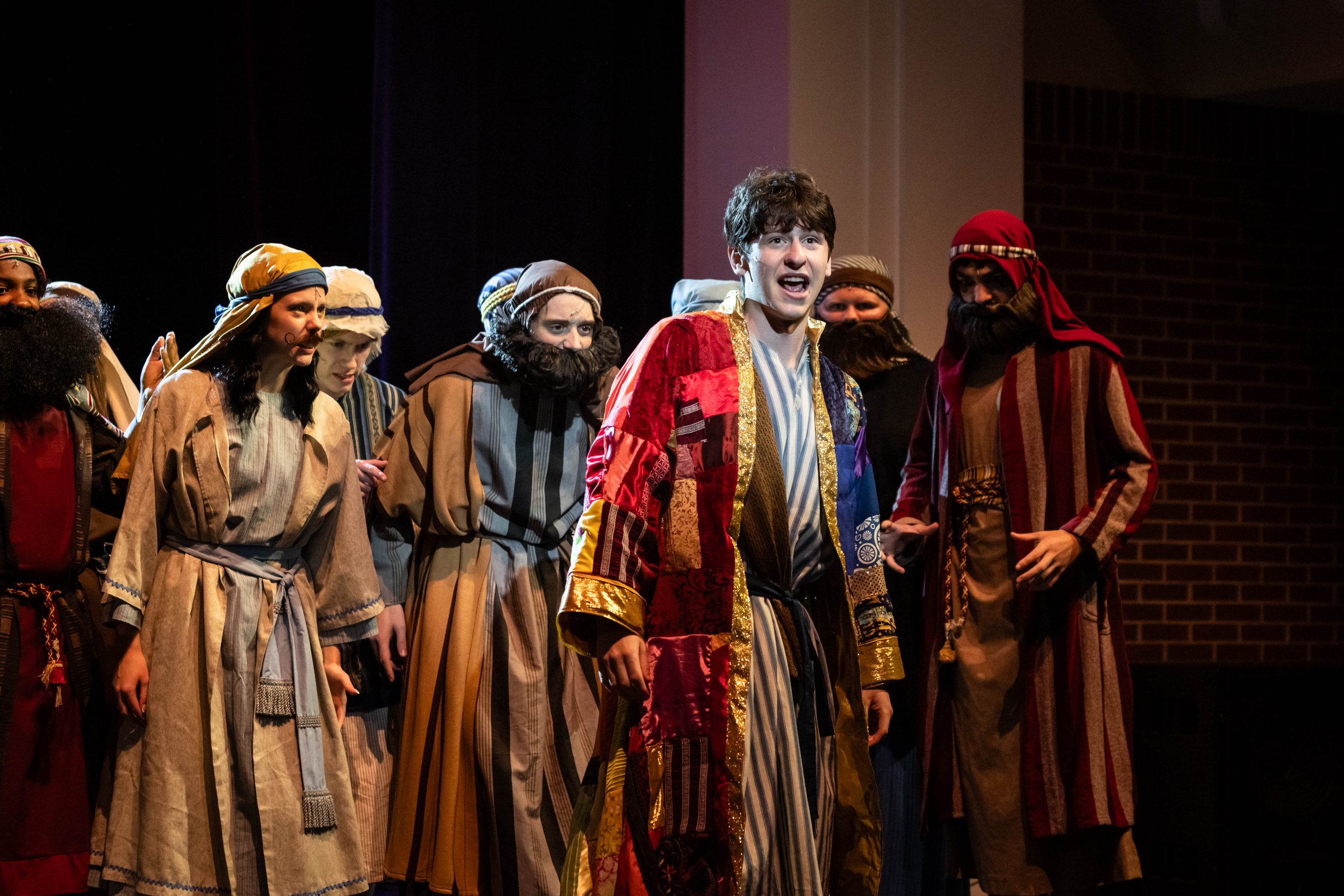 A group of actors on stage dressed as biblical characters, likely from a nativity play, with a young man in the center wearing a colorful, ornate robe and a gold trim, singing or speaking, while others dressed as shepherds and Wise Men stand around h