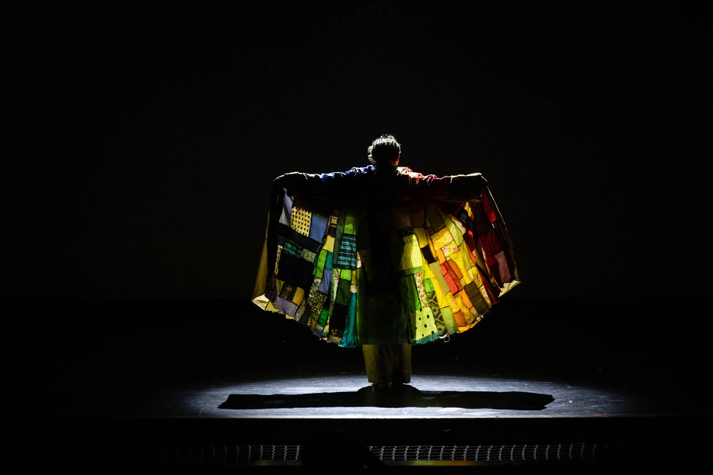 Person on stage dressed in a colorful patchwork costume, standing with arms outstretched, illuminated from below with dark background.