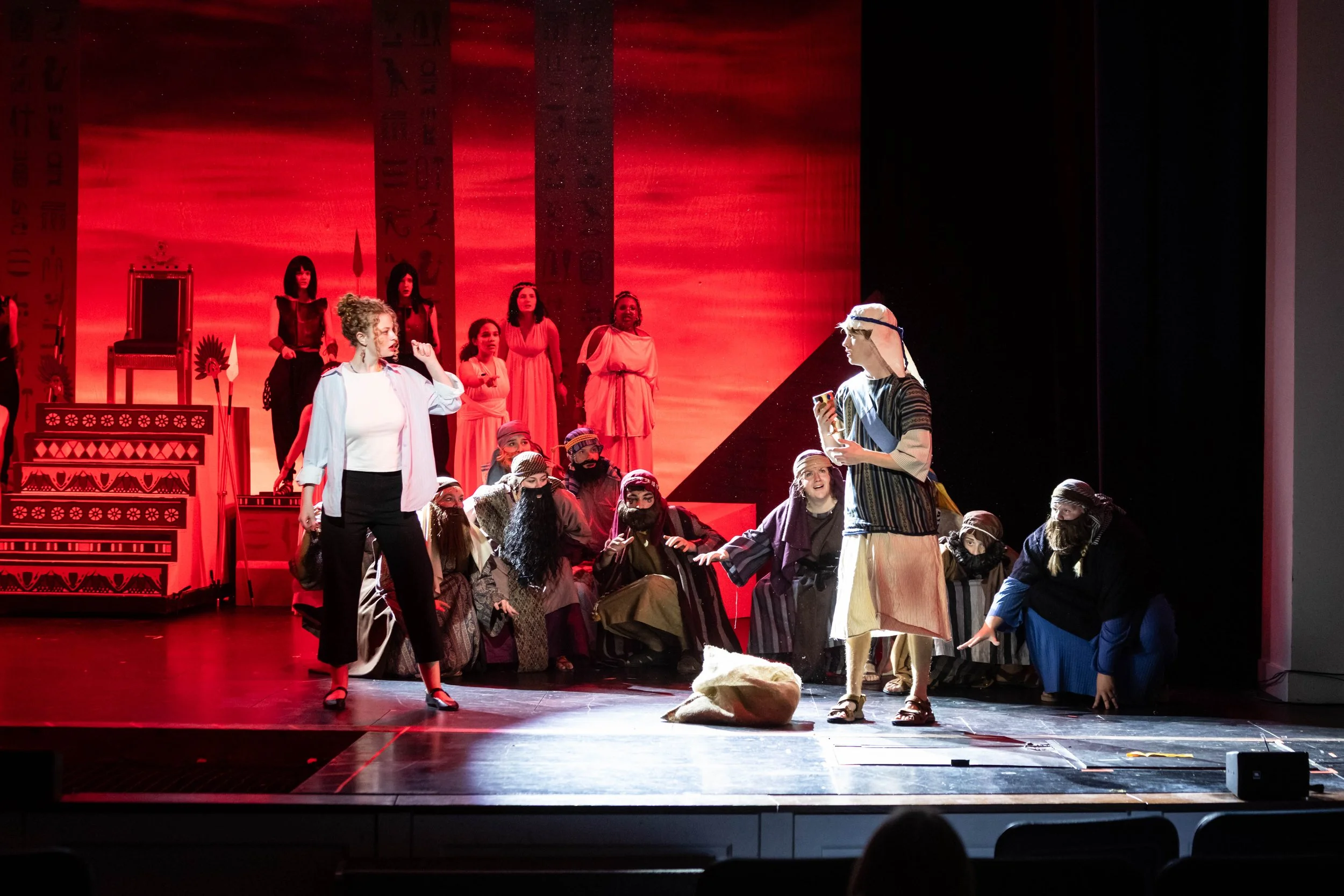 Theater stage with actors in costumes performing a play, featuring a woman in a striped dress and headband, and a group of actors in long robes and head coverings, with a red backdrop resembling ancient Egyptian hieroglyphs.