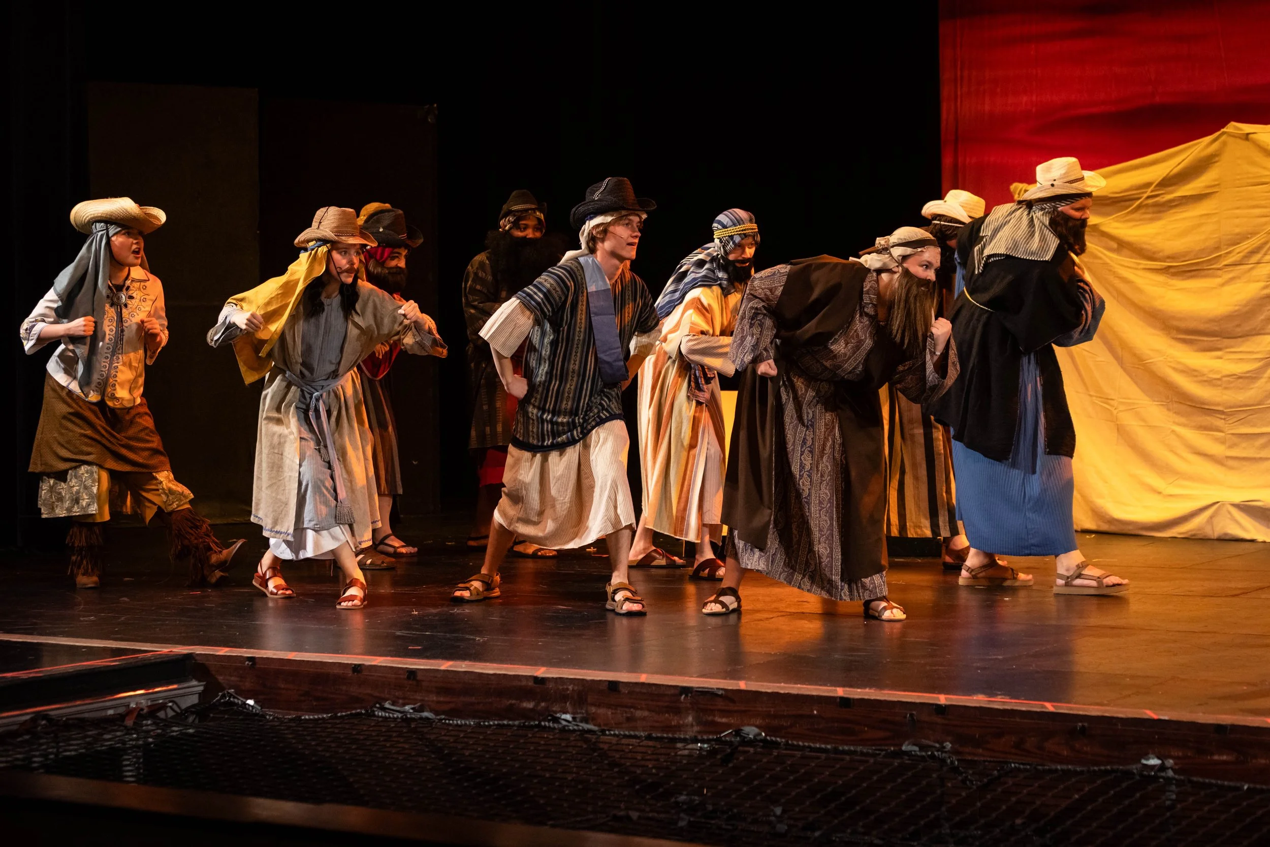 Actors dressed as biblical characters, including women in long robes and headscarves and men in costume with hats and beards, performing on stage in a play or religious reenactment.